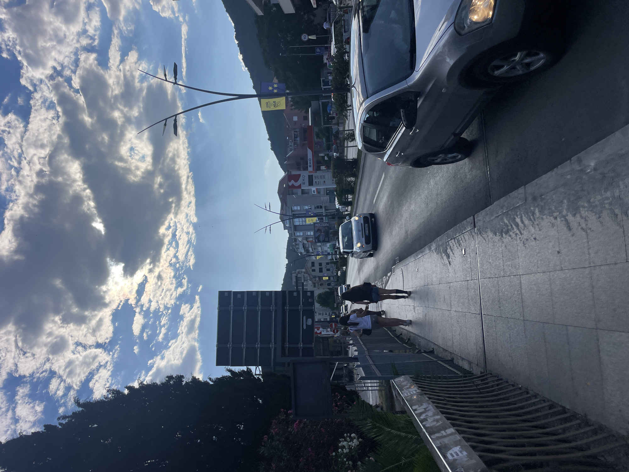 The image depicts a street scene in Budva, Montenegro, under a cloudy sky with sunlight breaking through. In the foreground, a silver car drives on the right side of the road. To the left, a sidewalk runs alongside the road, bordered by a metal fence. Two young women, dressed casually in shorts and t-shirts, walk away from the camera on the sidewalk. Behind them, a large, dark rectangular billboard is visible. Further down the street, buildings, including what appears to be a gas station with "EKO" branding, line the road. Streetlights with distinctive curved arms are positioned along the street. In the background, hills covered in vegetation rise above the town. The overall scene suggests a late afternoon or early evening setting in an urban coastal area.