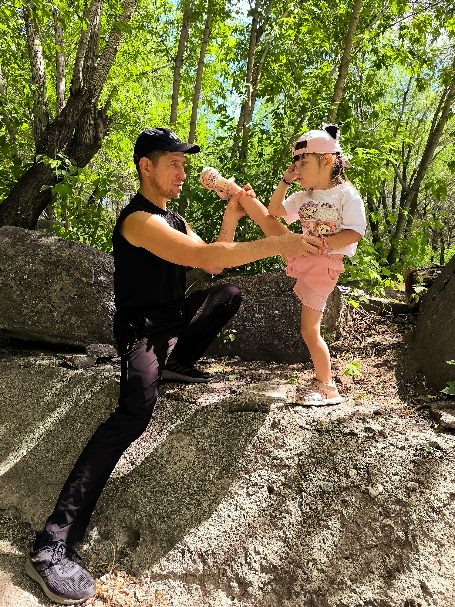 In a sun-dappled forest setting, a man and a young girl are engaged in a martial arts practice. The man, wearing a black sleeveless shirt, black pants, and a black baseball cap, is kneeling on a large rock. He is holding the girl's leg as she extends it upwards, demonstrating a kick.
The girl, dressed in a white t-shirt and pink shorts, wears a pink baseball cap with cat ears. She has a focused expression as she practices her kick. She is also wearing light pink sandals.
The environment is lush with green trees and foliage, suggesting a warm, likely summer day. Large rocks are scattered throughout the scene, forming part of the uneven terrain. The lighting indicates it is daytime, with sunlight filtering through the leaves and casting shadows.
Visible text on the girl's t-shirt reads: "HUNTRUNX".