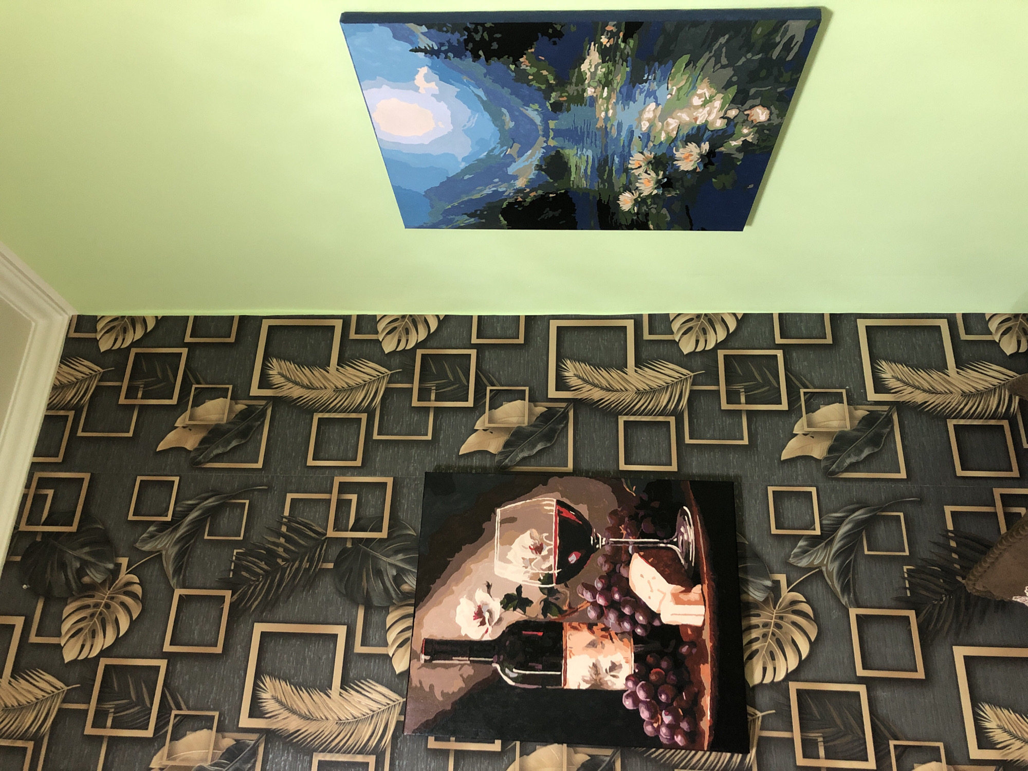 This is a medium-shot, eye-level photograph of two framed paintings hanging on a wall in a room, likely a living room or hallway. The wall on the left features a dark gray wallpaper with a repeating pattern of gold-colored squares and stylized leaves in shades of gold and dark green. The wall on the right is painted a light, pastel green.
The painting on the left depicts a still life of a bottle of red wine, a glass filled with red wine, a bunch of dark red grapes, and a wedge of cheese on a wooden surface. The style of this painting appears to be a "paint by numbers" artwork, given the distinct blocks of color and visible outlines.
The painting on the right is a landscape scene featuring a body of water, possibly a lake or river, with lily pads and flowers. Mountains and trees are visible in the background under a bright, stylized sky. This painting also exhibits characteristics of a "paint by numbers" artwork.
The lighting appears to be artificial, creating some reflections on the paintings. The perspective suggests the photograph was taken from an angle, with the camera looking towards a corner where the two walls meet. There are no people visible in the frame. The image does not contain any information that would place it in Safonovo, Russia.