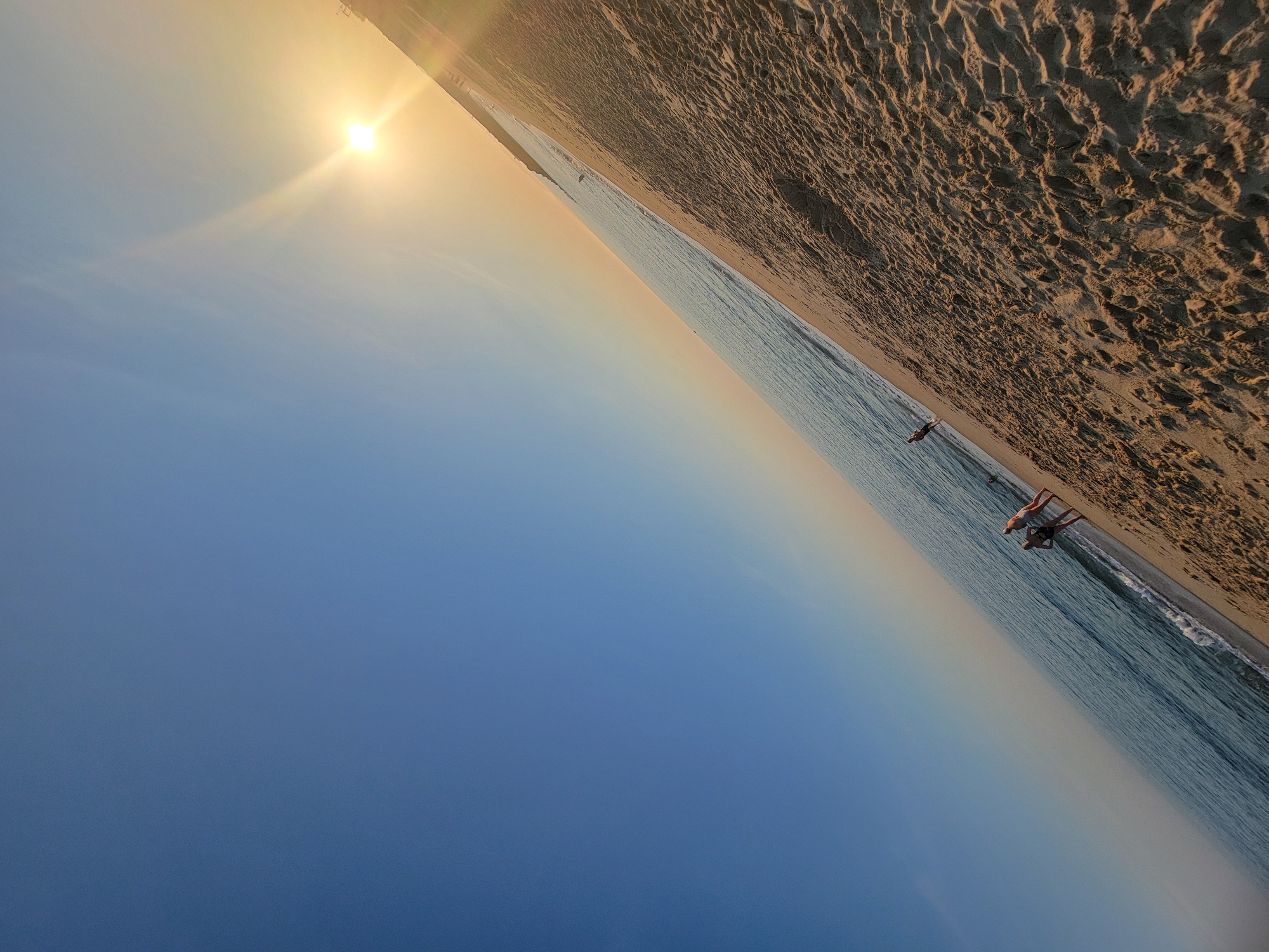 The image depicts a beach scene in Quarteira, Portugal, during what appears to be sunset or sunrise. The sun, a bright orb with visible rays, is low in the sky, casting a warm glow and creating a soft gradient in the atmosphere from a pale yellow near the horizon to a deep blue overhead. The ocean stretches out, with gentle waves lapping at the shore. The beach itself is sandy, with visible textures from the sand.
Several people are present, scattered along the shoreline. One person is in the water, appearing to be swimming or wading. Further up the beach, two individuals are standing, one in swimwear, suggesting they are enjoying the beach. The overall mood seems peaceful and serene, characteristic of a quiet moment by the sea. The composition is angled, with the sky occupying the left two-thirds of the frame and the beach and ocean to the right. There is no visible text in the image.