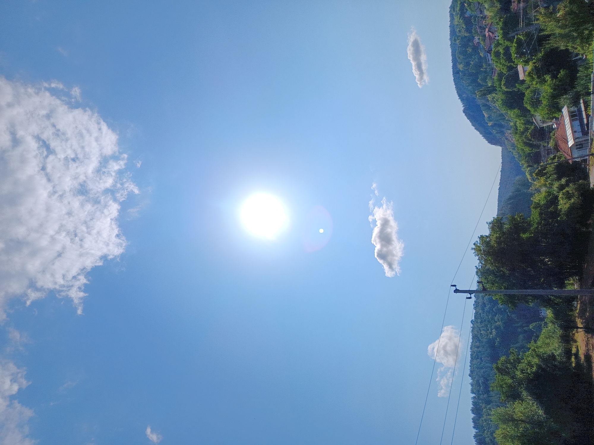 The image depicts a bright, sunny day in Kralev Dol, Bulgaria, with a clear blue sky dominating the upper half of the frame. The sun is high in the sky, appearing as a bright, almost blinding orb with a noticeable lens flare and a smaller, circular reflection. Several scattered white clouds drift across the sky, with a larger cluster near the top and smaller ones lower down.
The lower portion of the image shows a verdant landscape. Rolling hills covered in dense green trees recede into the background. A village or settlement is visible on the slopes of the hills, with numerous houses featuring red-tiled roofs interspersed among the foliage. A power line with insulators and wires stretches horizontally across the mid-ground, supported by a utility pole on the right side of the frame. The foreground consists of a dry, grassy area with scattered trees and some patchy vegetation, suggesting a rural or semi-rural setting. The overall impression is one of a warm, summer day in a picturesque Bulgarian countryside.