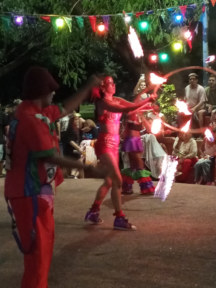 Two fire performers entertain an outdoor crowd under strings of colorful lights. The woman, wearing a sparkly pink costume, expertly spins flaming objects. Her male partner, dressed in red with patchwork details, gestures along with her performance. Spectators are seated and standing, watching the show.