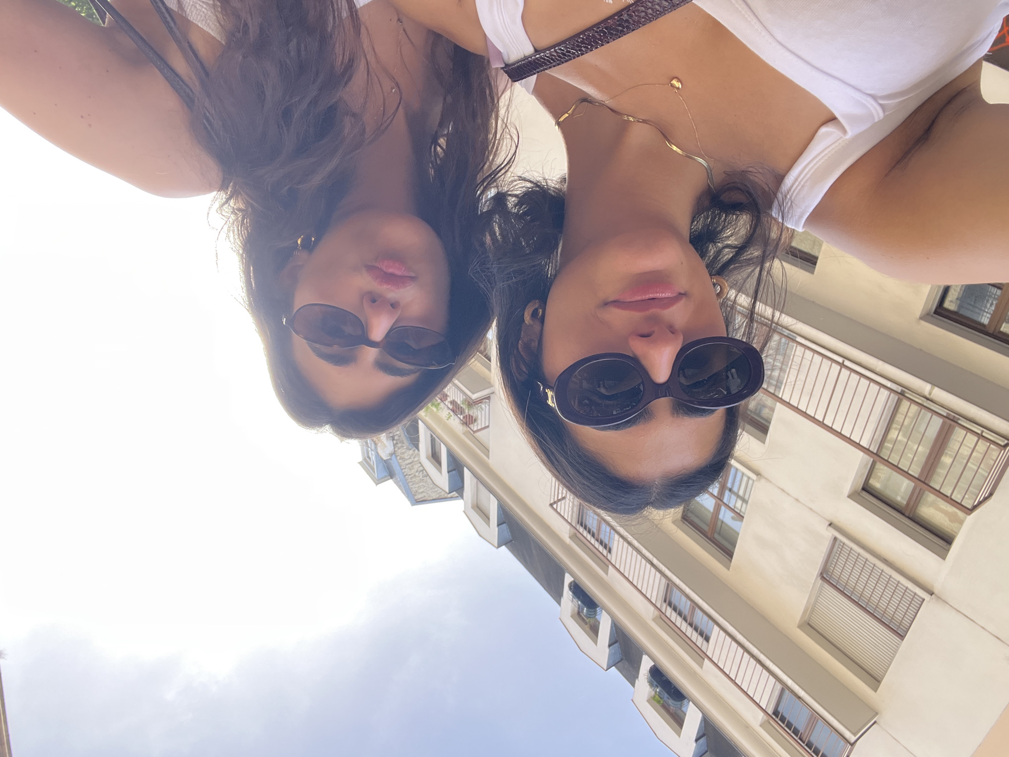 Two women are posing for a selfie outdoors on a sunny day. They are wearing sunglasses and white tank tops. One woman has a brown purse strap over her shoulder and a gold necklace. The background shows a building with several windows and balconies. The scene is likely in Paris, France, based on the architectural style of the building.