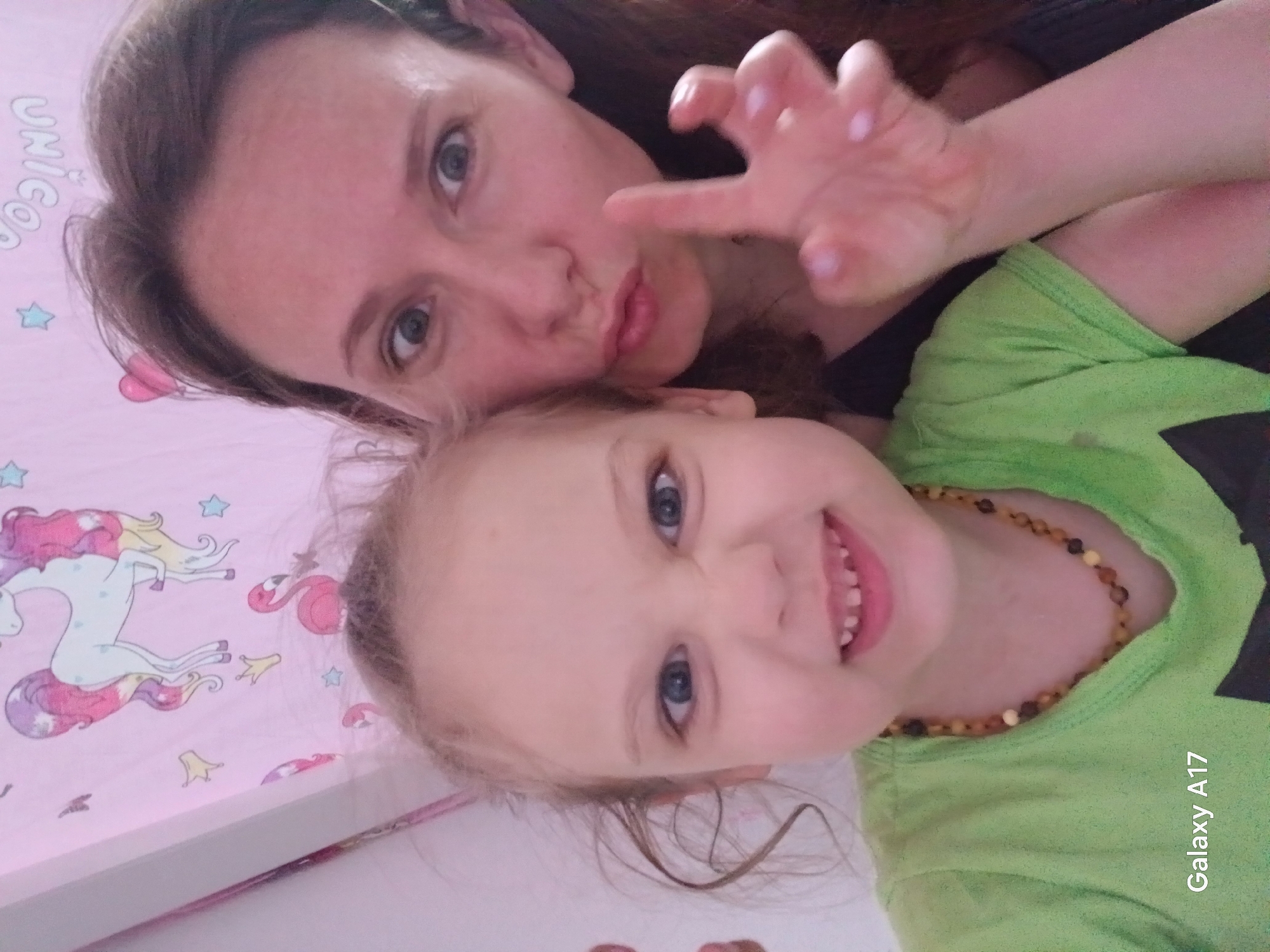 A selfie shows a woman and a young girl in close proximity. The woman, with light skin and brown hair, is making a playful "claw" gesture with one hand and pursing her lips in a kiss. The girl, also with light skin, blonde hair, and blue eyes, is smiling broadly, revealing her teeth. She is wearing a green shirt and a beaded necklace. They are lying on a pink surface decorated with cartoon unicorns and stars. A "Galaxy A17" watermark is visible in the lower right corner. There is no evidence to indicate that the image was taken in Yekaterinburg, Russia.