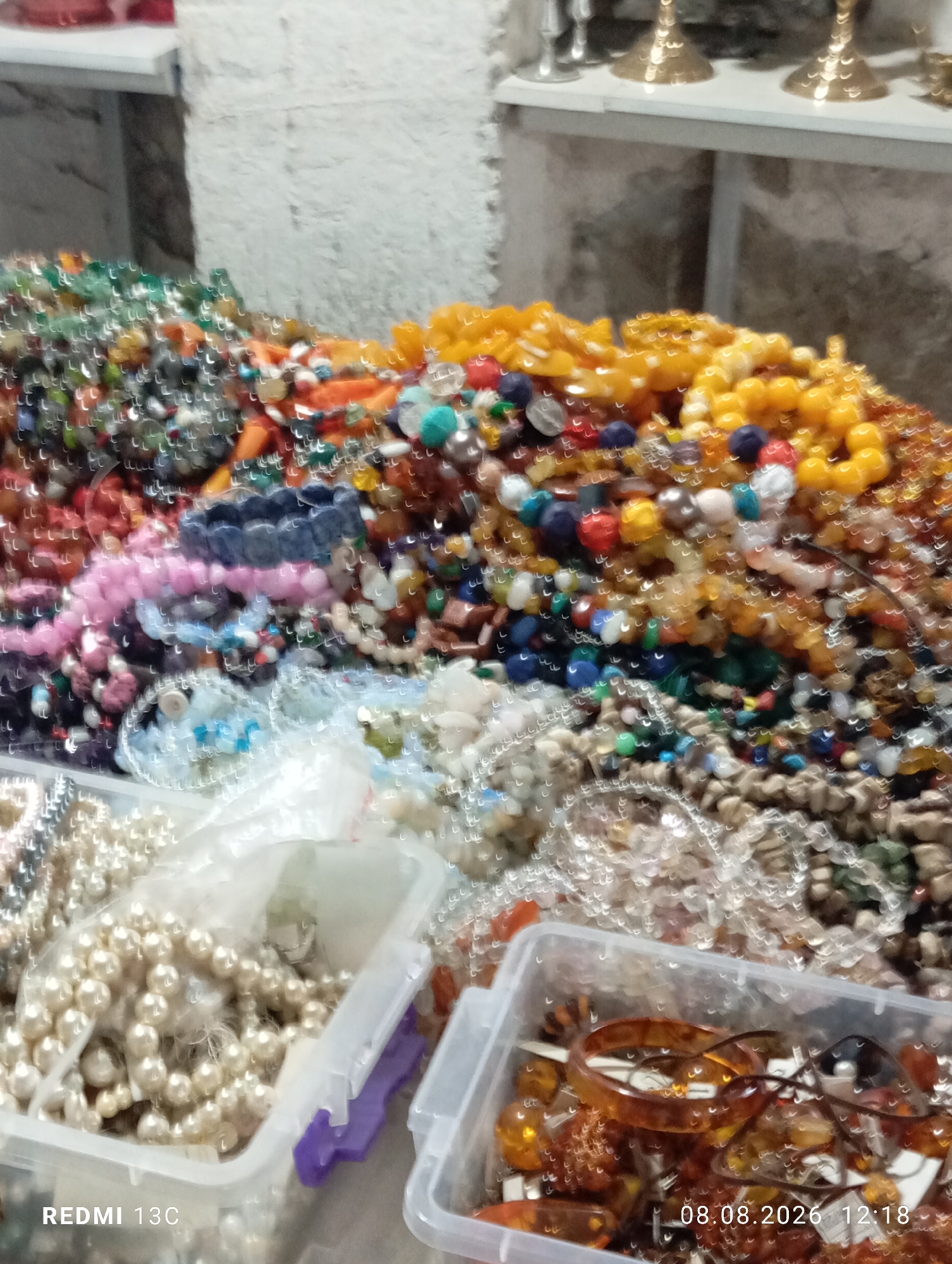 The image shows a close-up, slightly out-of-focus view of a variety of colorful beads and jewelry items displayed in transparent plastic containers. The beads are of various sizes, shapes, and colors, including pearls, amber-colored pieces, and multi-colored gemstones. Some of the jewelry appears to be strung into necklaces or bracelets. In the background, there is a textured wall and what looks like shelves with decorative items. The overall impression is of a market stall or a shop specializing in beads and accessories. The timestamp "08.08.2026 12:18" and the text "REDMI 13C" are superimposed on the image, indicating the date and time the photo was taken, and possibly the device used. Based on the provided context that this image is located in Tashkent, Uzbekistan, this scene likely depicts a stall in a bazaar or a jewelry shop within the city. There are no people visible in the image.