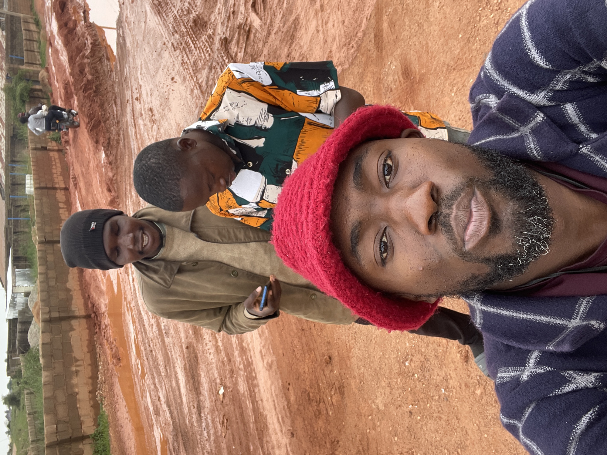 In the foreground, a man wearing a red knitted hat and a dark blue and gray checkered jacket looks directly at the camera, puckering his lips. Behind him, to his left, another man in a black beanie and a tan jacket holds a blue smartphone. To the right of the man with the beanie, a third man in a colorful, patterned shirt looks down. The background shows a muddy, unpaved road with puddles of water, likely in an undeveloped area of Jos, Nigeria. A brick wall runs along the back of the scene, with some scattered debris and buildings in the distance. A motorcycle with two riders is visible further down the road, suggesting ongoing activity in the area. The overall impression is a candid moment captured outdoors in a rural or developing urban setting.
