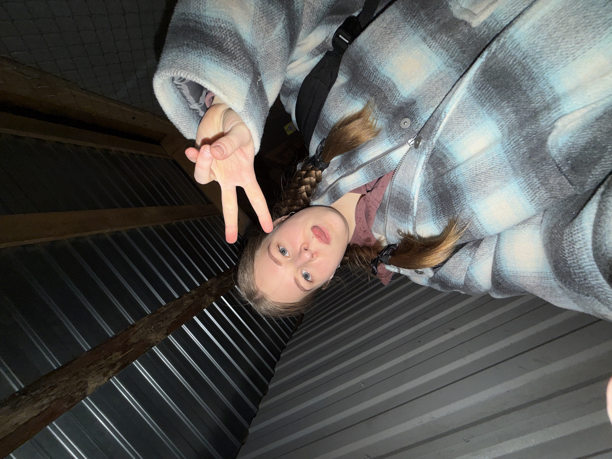The image is taken at an angle from below, showing a young woman in a gray and blue plaid jacket. She is making a peace sign with her left hand. Her hair is in braids, and she is looking down at the camera with her mouth slightly open. The background appears to be the underside of a metal roof with wooden beams. The location is assumed to be in the city of Perm, Russia, due to the image's origin.