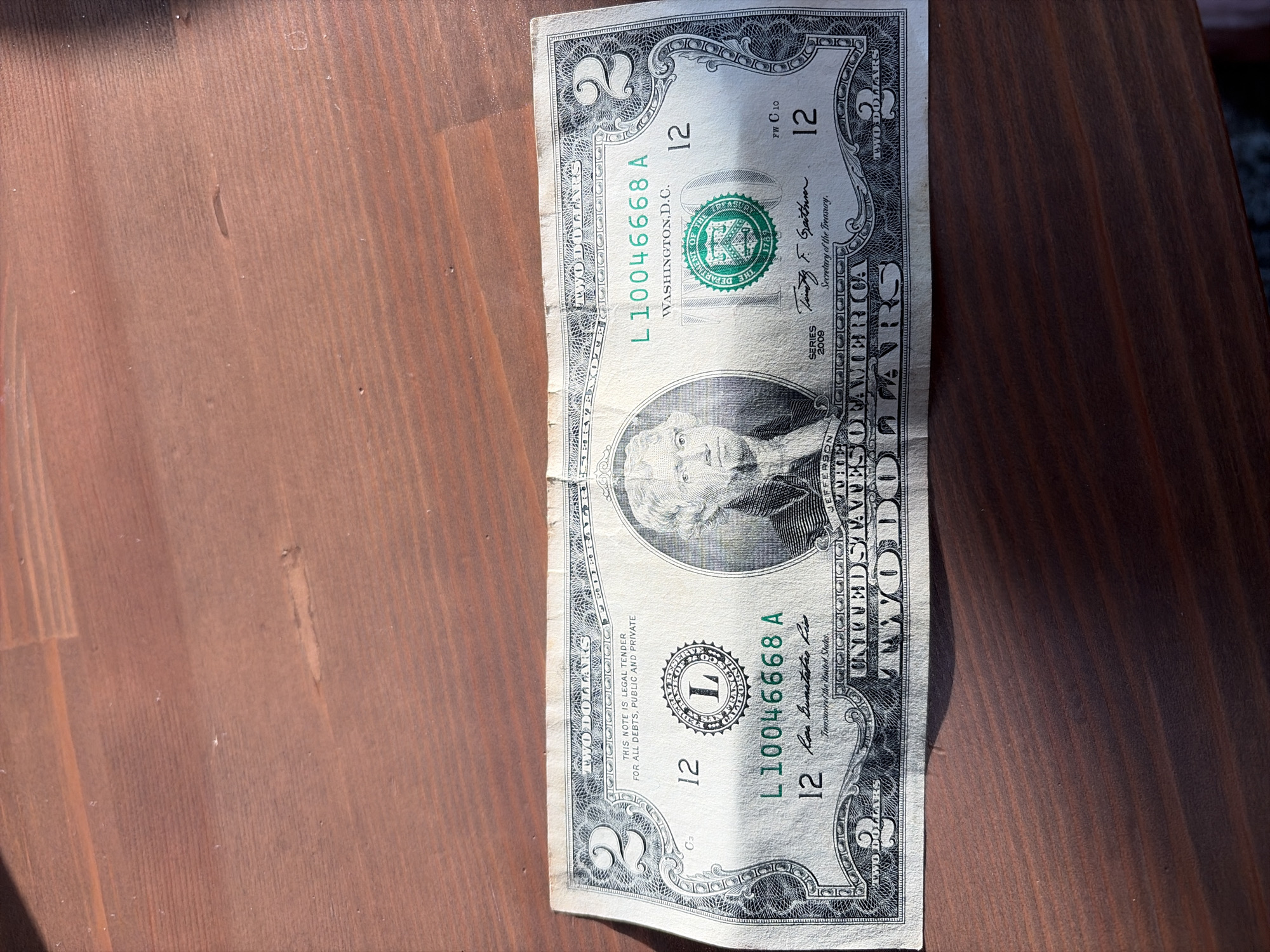 This media file features a close-up, overhead view of a U.S. two-dollar bill placed on a dark wooden surface. The bill is positioned vertically, with the top of the bill slightly blurred. The wooden surface appears to be made of planks with a visible grain.
The bill itself shows the portrait of Thomas Jefferson on the left side and a scene of the signing of the Declaration of Independence on the back (not visible in this particular image, which focuses on the front). Visible text on the bill includes "UNITED STATES OF AMERICA," "TWO DOLLARS," "WASHINGTON, D.C.," the serial number "L10046668 A" appearing twice, the series year "2009," and signatures of the Secretary of the Treasury and Treasurer. The Federal Reserve seal and the U.S. Treasury seal are also clearly visible.
The lighting suggests it is daytime, casting a slight shadow to the right of the bill. There are no people or animals present, and the activity appears to be static, simply showcasing the currency. The location context provided is Yekaterinburg, Russia, but there are no specific visual cues in the image to confirm this location; the wooden surface and the U.S. dollar bill are generic.