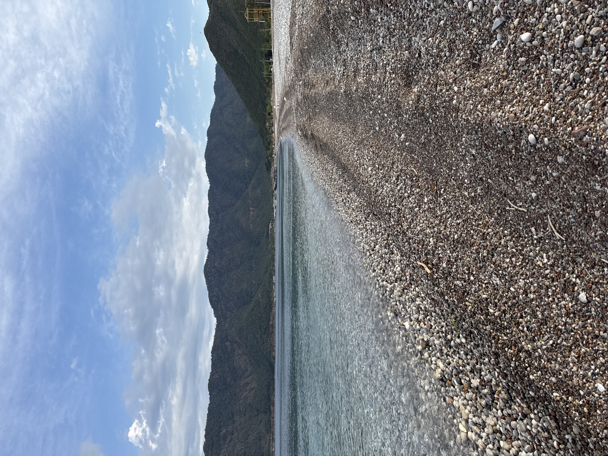 The media file captures a serene coastal landscape in Kumluca, Turkey, under a bright, partly cloudy sky. The image is composed of three distinct horizontal sections: a pebble beach in the foreground, calm water in the midground, and large mountains in the background.
In the immediate foreground, a wide expanse of a pebble beach stretches across the right side of the frame, comprising countless small, irregularly shaped stones in shades of grey, brown, and white. These pebbles transition towards the water, where they become wet and darker. The midground features clear, tranquil water, likely the sea or a large lake, showing gentle ripples and a smooth surface that reflects the sky faintly.
Dominating the left side of the scene are massive, dark green mountains, covered in dense vegetation, rising steeply from the water's edge. Their peaks extend into the upper portion of the frame. In the far distance, nestled against the base or slope of the mountains, some faint structures, including a yellow scaffolding-like element, are discernible, hinting at distant human presence. The sky above is a bright blue, dotted with scattered white cumulus and cirrus clouds, suggesting a pleasant, sunny day, likely late morning or early afternoon.
There are no people or specific activities visible, emphasizing the natural and untouched beauty of the location. No visible text is present in the image. The overall impression is one of peace and natural grandeur.