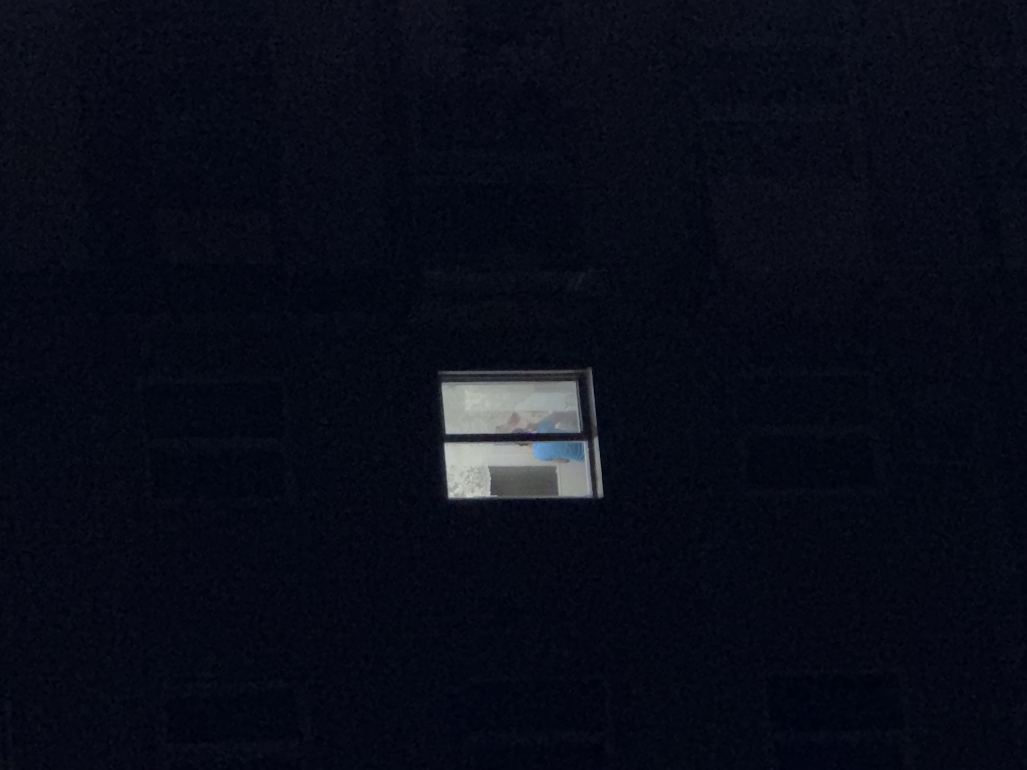 An exterior, medium-shot, eye-level view shows a building facade at night. The facade is dark, and the only visible light source is a single, illuminated window in the center of the frame. Inside the window, two figures are visible. One figure, wearing a blue shirt, is seated. The other figure is standing and appears to be interacting with the seated figure. The interior of the room is dimly lit, with patterned curtains visible behind the figures. The scene suggests an urban environment, likely residential, given the building's appearance and the presence of an apartment window.
