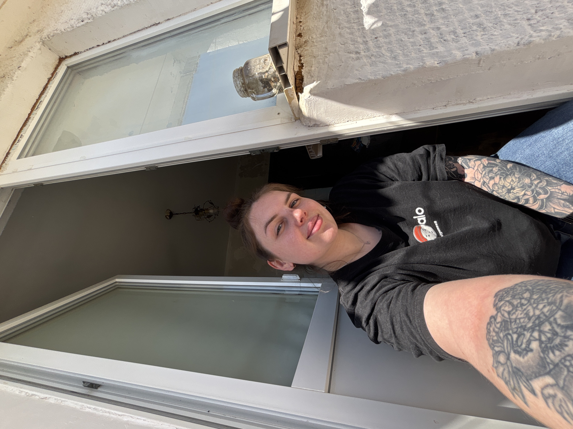 A person is leaning out of a window. The person is wearing a black t-shirt and has tattoos on their arms. The window is white and has a clear glass pane. The location is Chornomorskoe, Ukraine. The scene appears to be inside a building, looking out through the window. The wall around the window is white.