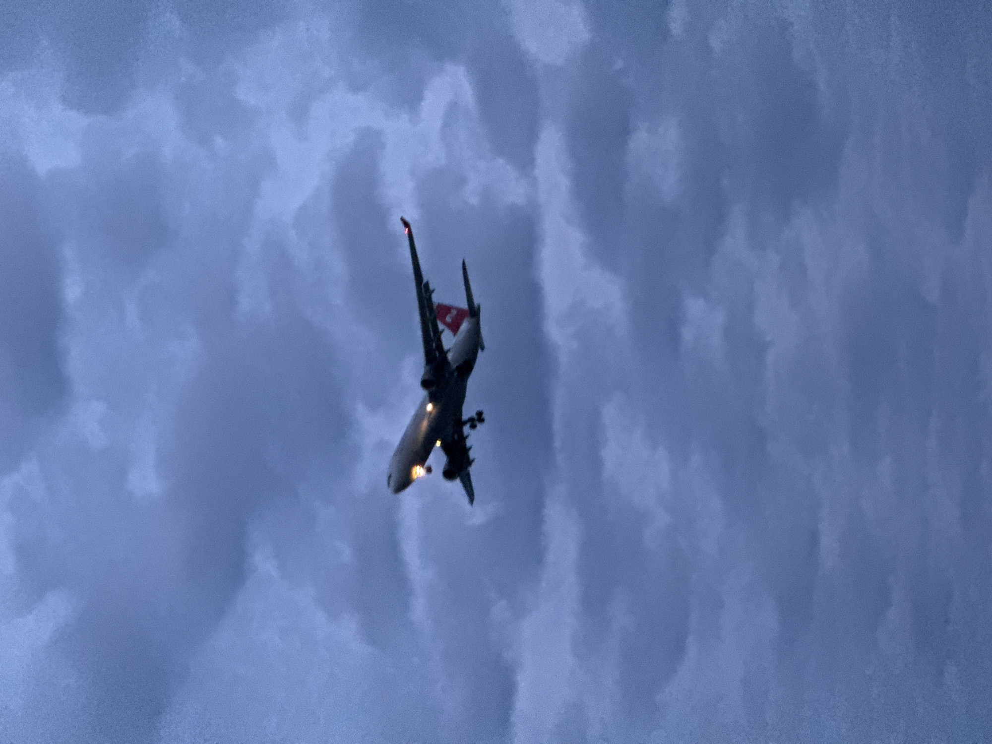 An airplane is captured in mid-flight against a heavily overcast, blue-grey sky. The aircraft is viewed from below, appearing to be either landing or taking off, as its landing gear is extended and multiple lights are illuminated on its underside. The plane's fuselage is white, and its vertical stabilizer features a distinct red color with a white emblem, resembling a flag, visible near the top. The sky is filled with soft, diffused clouds, indicating low light conditions such as dusk, dawn, or a deeply overcast day. The overall atmosphere is dim and muted. No specific text is discernible on the aircraft or in the cloudy sky. The scene is set in Sochi, Russia.