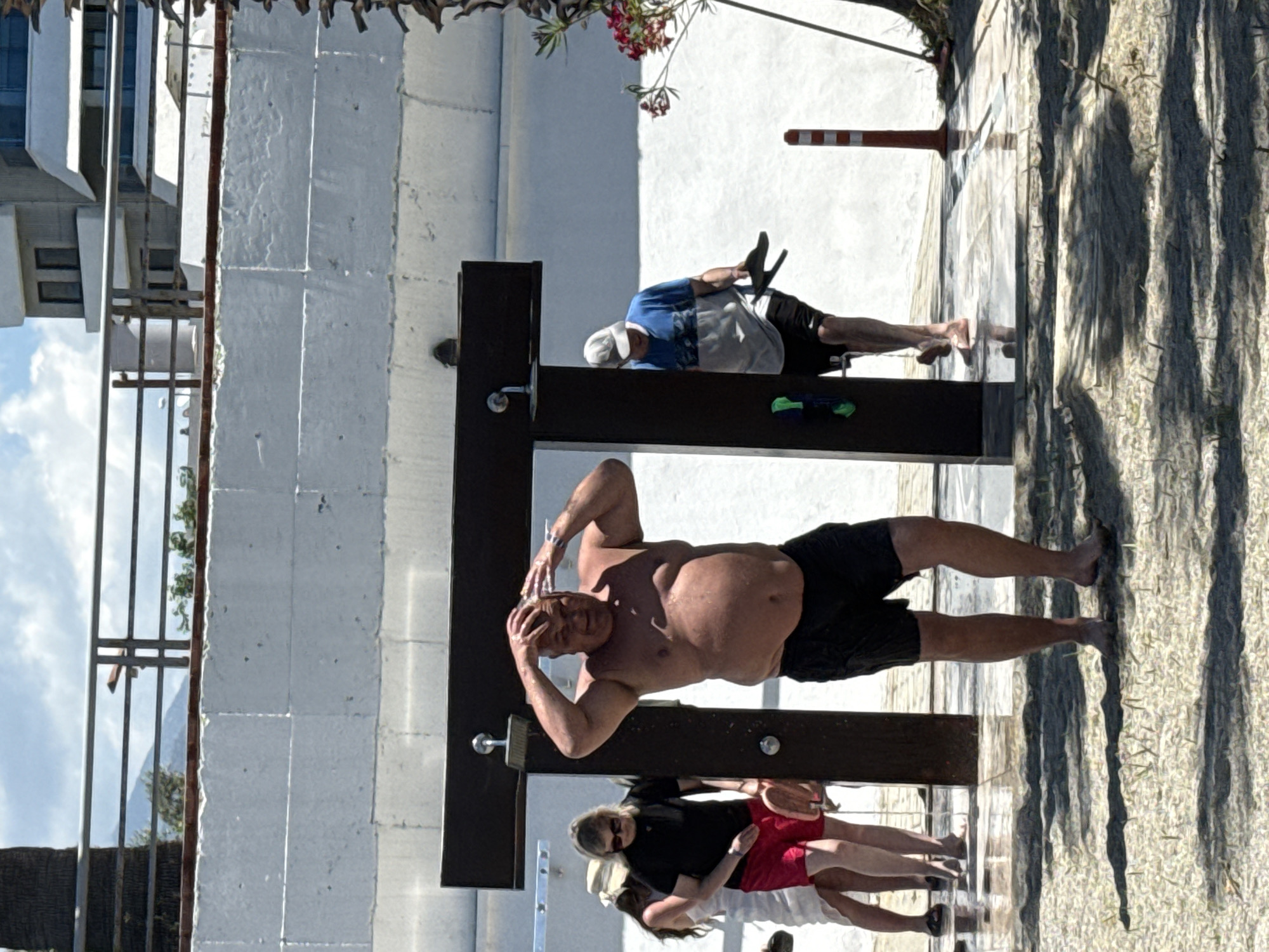 The image shows people using outdoor showers. A shirtless man is washing his hair at one shower, while another person in a blue shirt is at a different shower nearby. Other people are walking in the background, and shadows suggest a sunny day. The setting appears to be a public beach area, based on the nearby trees and foliage.