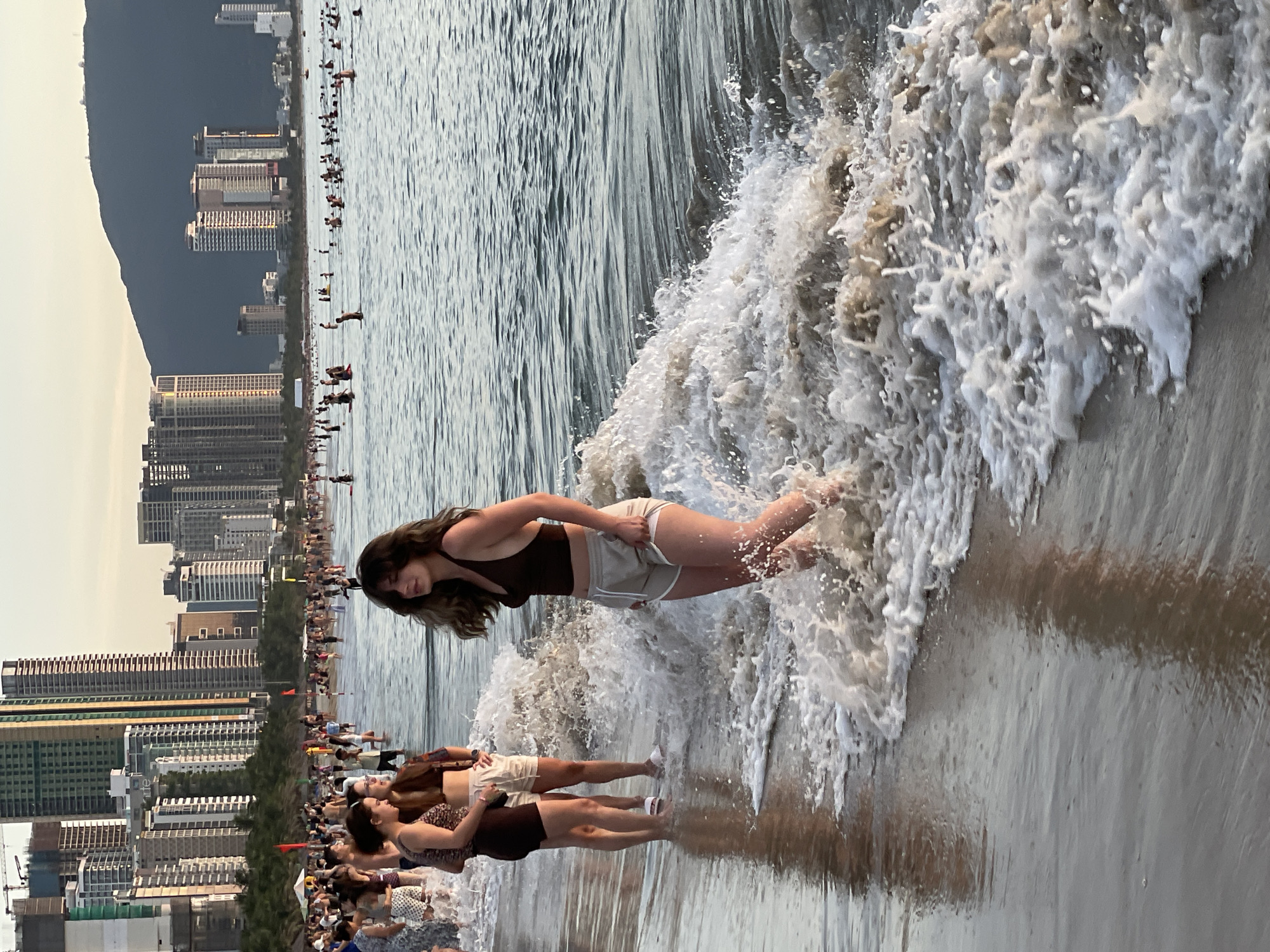 The image depicts a crowded beach scene in Da Nang, Vietnam, at sunset. In the foreground, a young woman in a dark brown bikini top and light-colored shorts stands in the shallow surf, her hair blowing in the breeze. To her left, two other young women stand in the water, one wearing a patterned bikini top and dark shorts, the other in a brown bikini top and light-colored shorts. They are looking towards the woman in the foreground.
In the midground, the ocean is populated with numerous swimmers, many of whom are partially submerged or wading in the water. Further back, the beach is lined with many people, some walking and others gathered in groups. The background features a dense cityscape with numerous high-rise buildings, some under construction, characteristic of Da Nang's urban development. A dark, mountainous landscape is visible in the far distance, partially obscured by the buildings. The sky is a soft, warm hue, indicating the time of day is near sunset. The overall atmosphere is one of a vibrant, popular tourist destination.