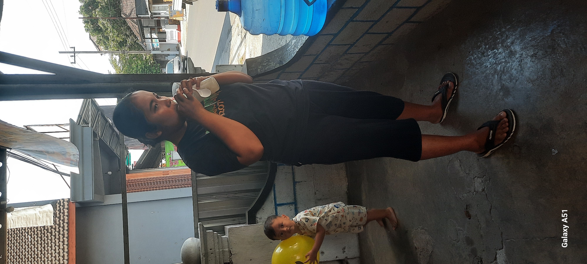 In Selopuro, Indonesia, a woman wearing a black t-shirt and black cropped pants, and flip-flops, stands and drinks from a clear plastic cup. Beside her, a young boy wearing a white patterned shirt and shorts, with bare feet, holds a large yellow balloon. The scene takes place outdoors, with a brick wall behind them and a large blue water jug to the woman's right. In the background, a street is visible with houses and trees, suggesting a residential or commercial area.