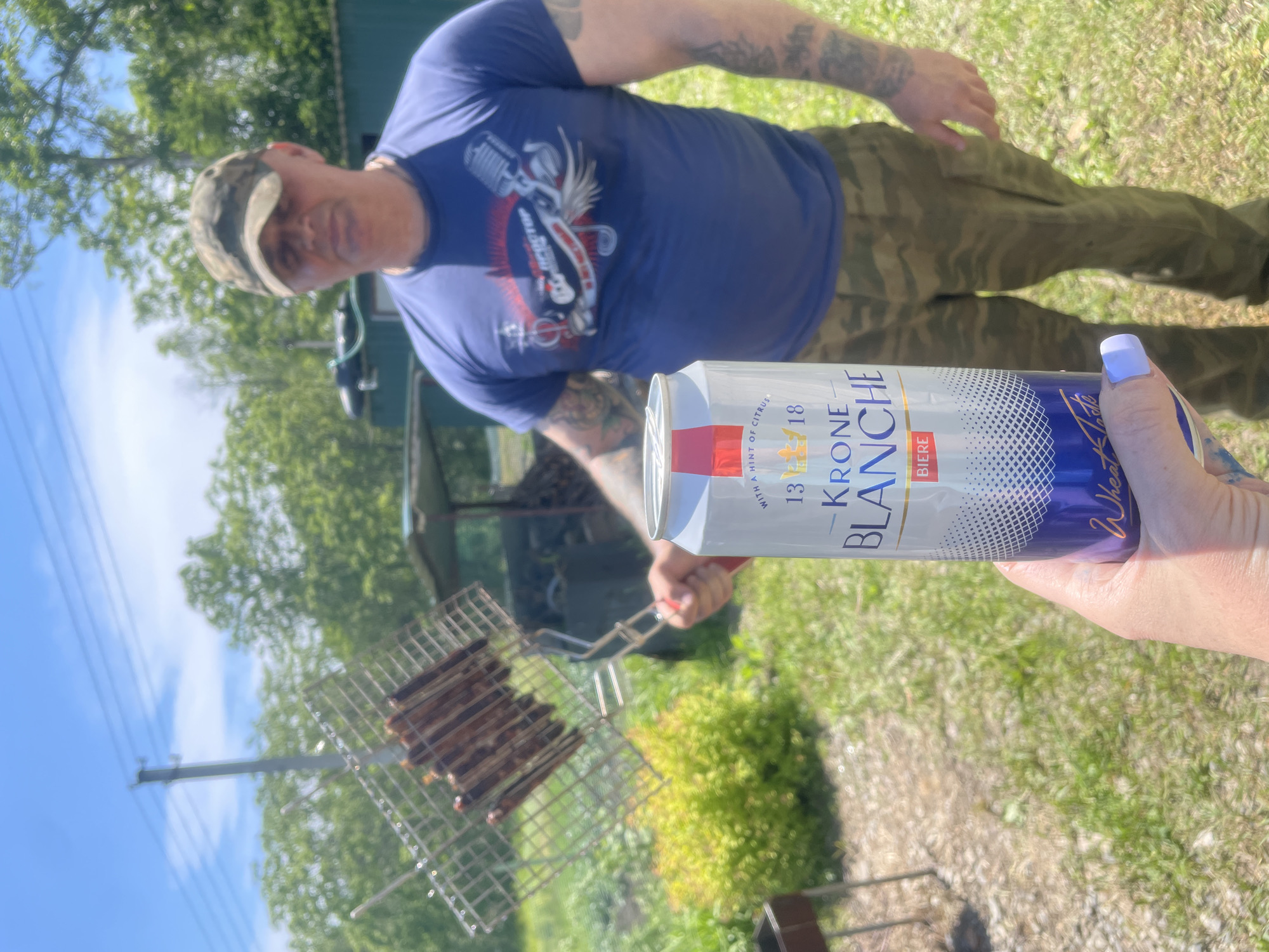 The image depicts an outdoor scene, likely in Yasnoe, Russia, with a person in the foreground holding a can of Krone Blanche beer. The can is white with blue and gold accents, and text indicating "BIERE" and "WITH A HINT OF CITRUS." Behind the beer can, a man wearing a camouflage baseball cap, a blue t-shirt with a graphic design, and camouflage pants stands in the middle ground. He is holding a metal grill basket filled with food, possibly sausages, over what appears to be a barbecue. The background shows a grassy area with trees and a green building, suggesting a rural or semi-rural setting. The lighting indicates it is daytime, likely sunny, with a clear blue sky.