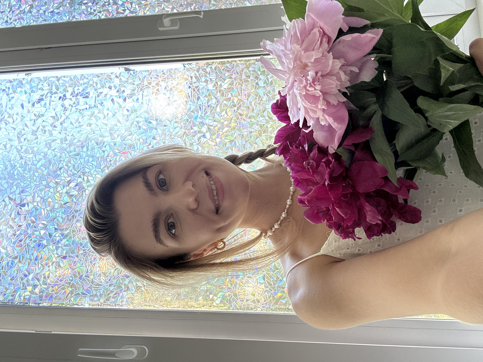 A young Caucasian woman with blonde hair styled in a braid and her hair down, is holding a bouquet of pink and magenta peonies in front of a window with a textured, multicolored film. She is wearing a white dress with a pearl necklace and hoop earrings. The woman is smiling at the camera, with her dimples visible. The background consists of a window with a stained-glass-like film that diffuses the sunlight, creating a vibrant, colorful effect. The overall scene suggests a bright, cheerful, and possibly indoor setting.