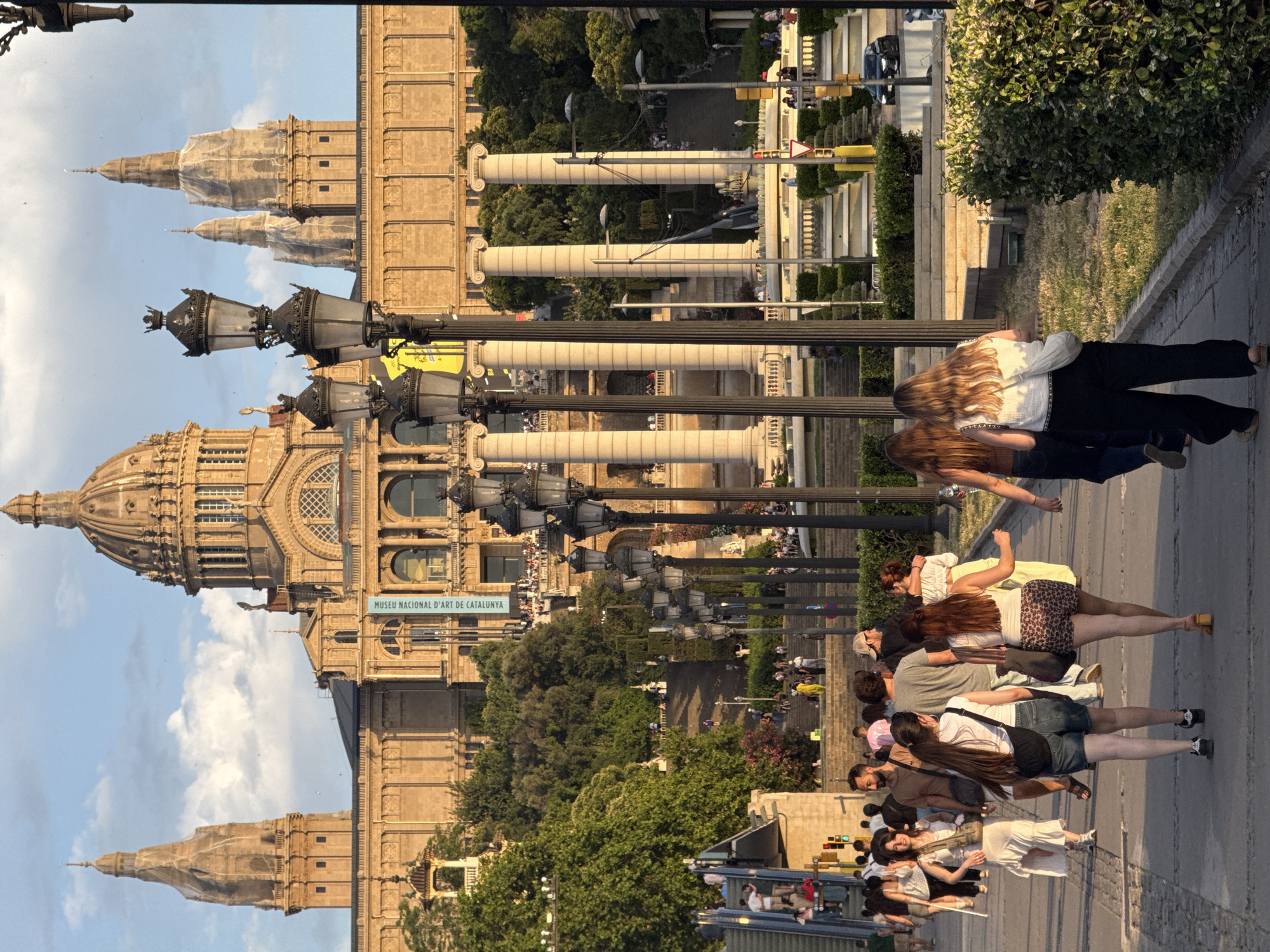 The image captures a scene in front of the Palau Nacional, located on Montjuïc hill in Barcelona, Spain. The grand building, a former palace and now home to the National Art Museum of Catalonia, dominates the background with its distinctive dome and towers. The late afternoon sun casts a warm glow on the building's facade.
In the foreground, a paved pathway is lined with people walking and milling about. Several street lamps with ornate black metalwork and glass casings stand along the pathway. A group of women are walking away from the camera, their backs to the viewer. One woman in the center wears denim shorts and a white top, while the woman to her right has on leopard-print shorts and a white tank top. Another woman on the left, facing slightly towards the camera, wears a light-colored dress. Men and women of various ages are also visible in the middle ground, some walking up a large set of stairs that leads to the Palau Nacional. Lush green trees and manicured hedges are interspersed throughout the scene, adding a natural element to the urban landscape. In the far background, traffic is visible on a road.