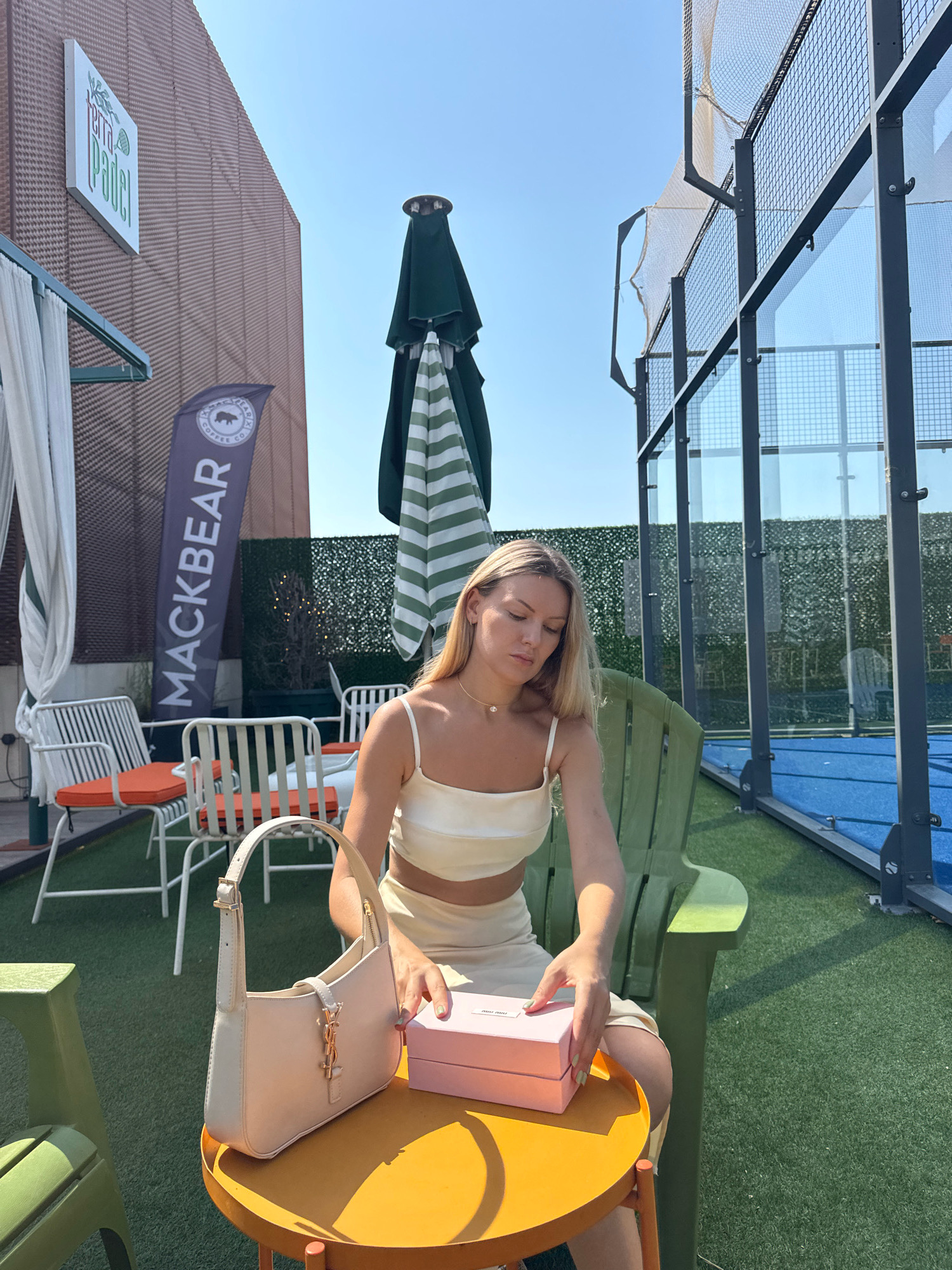A blonde woman wearing a cream-colored outfit sits in a green chair next to a small yellow table at a paddle tennis court. She is looking down at a pink box on the table. A cream-colored purse is also on the table. In the background, there are paddle tennis nets and courts, along with outdoor furniture and umbrellas. A banner with "MACKBEAR" and a logo featuring a bear is visible on the left side of the image. A sign with "terra padel" is mounted on a building. The scene appears to be an outdoor recreation area in the city of Antalya, Turkey.