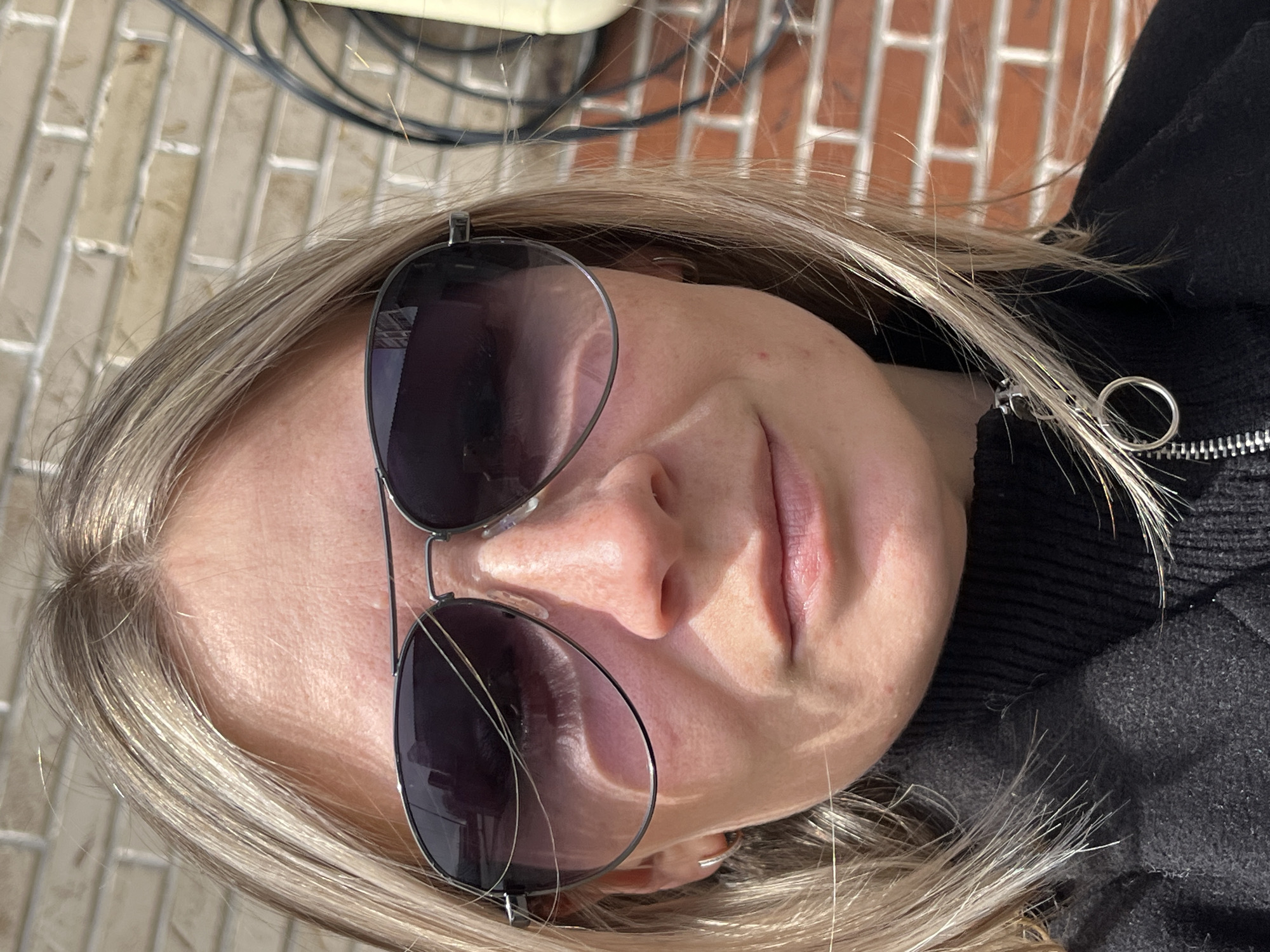 A close-up portrait-style image shows a person, likely a woman, outdoors on a sunny day in Chaikovsky, Russia. Her head is tilted, with her face partially visible, illuminated by bright natural light. She has light-colored, possibly blonde or light brown, hair, with strands framing her face and falling over her shoulders.
She is wearing aviator-style sunglasses with dark lenses, which obscure her eyes. Her lips are slightly parted, suggesting a neutral or subtly contemplative expression. She is dressed in a dark, possibly black or dark gray, ribbed or knitted garment, which appears to be a sweater or jacket, with a distinctive silver ring zipper pull visible on the right side of the frame.
The background includes a reddish-brown brick wall, suggesting an urban or suburban outdoor setting. Some dark cables or wires are visible in the upper background. The bright sunlight creates highlights on her hair and skin, indicating daytime with clear weather. There is no visible text in the image.
