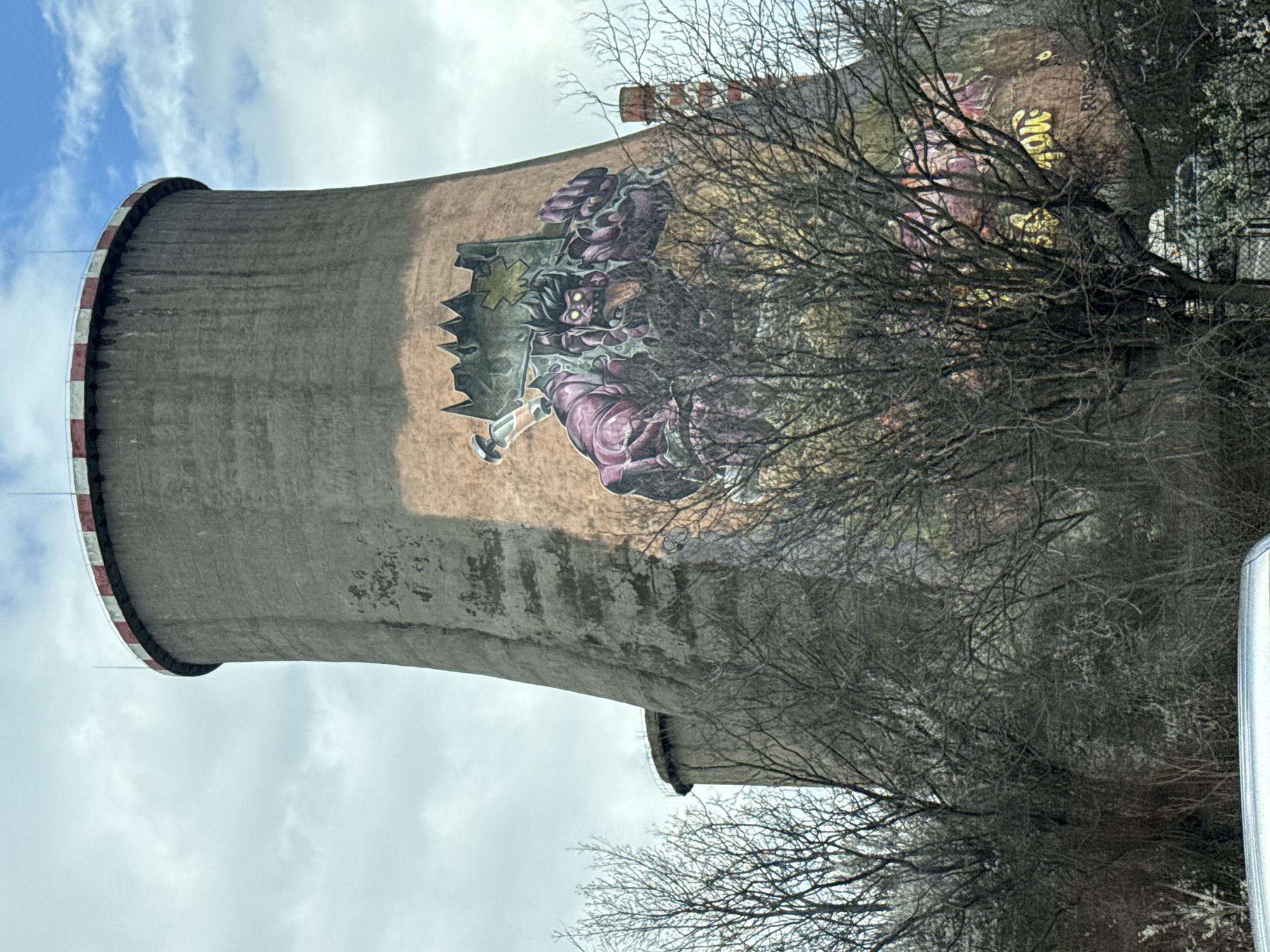 A cooling tower adorned with a mural is centered in the frame. A figure wearing a crown is depicted on the tower's surface, partially obscured by bare branches of a tree in the foreground. The location is the former factory near Zemlyane Depot in Sofia, Bulgaria, where towers were painted in 2017. The sky is cloudy with patches of blue.