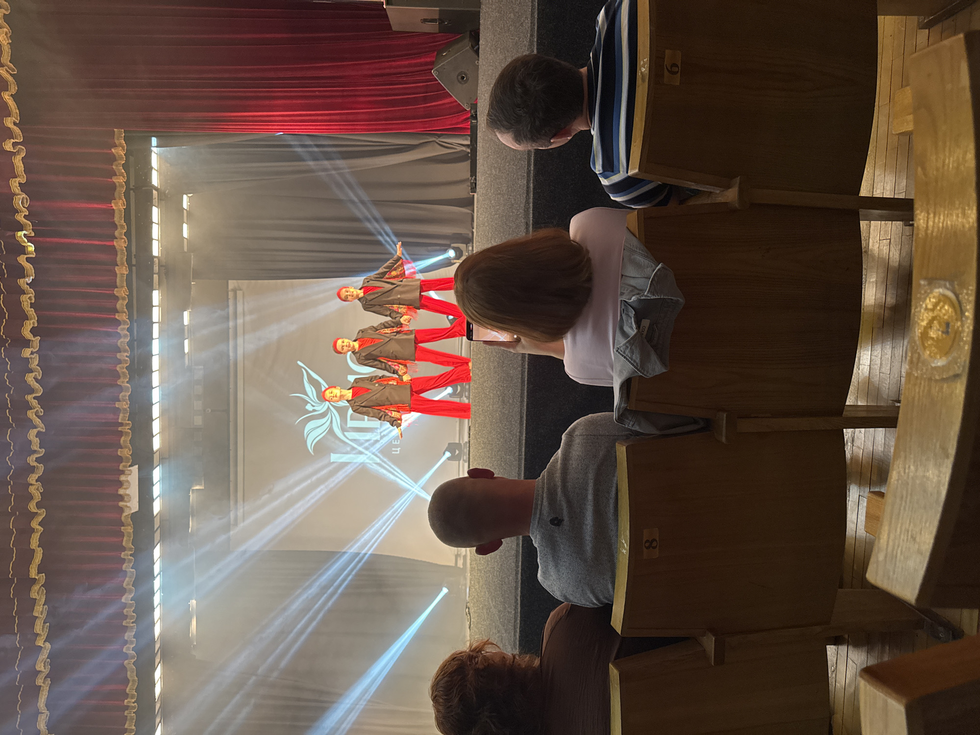 The image shows an indoor performance on a stage with three young performers in matching red pants and dark jackets with red fringe. They are illuminated by bright white stage lights. In the foreground, an audience watches from wooden chairs. The scene is set in the city of Sochi, Russia, likely within a community center or cultural facility. The performers appear to be engaged in a choreographed dance routine. The audience members are seated, with one person holding up a phone to record. The overall atmosphere suggests a local event or competition.