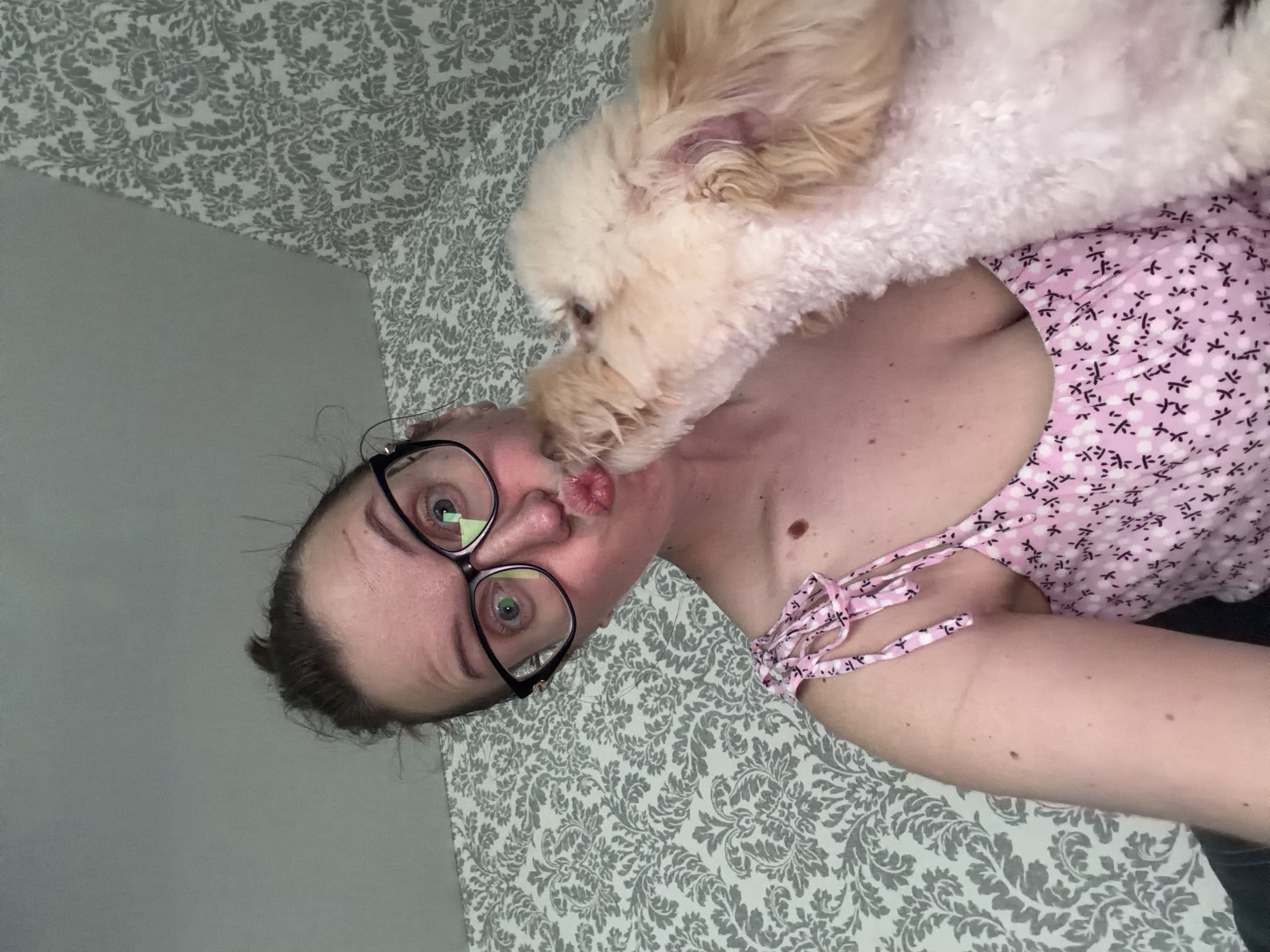 A woman with her hair pulled up in a bun and wearing glasses is puckering her lips as a light-colored, fluffy dog licks her face. She is wearing a pink floral-patterned top with tie straps. The background consists of patterned wallpaper. The image does not contain information about the location in Bila Tserkva, Ukraine.