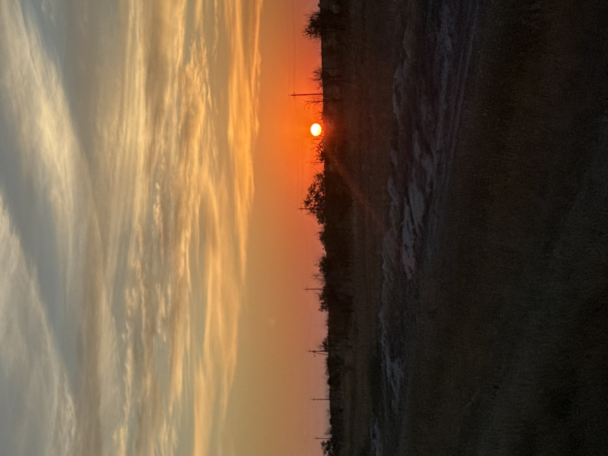 The image depicts a sunset scene in the village of Pugachëv, Kazakhstan. The sun, a bright orange orb, is low on the horizon, casting a warm glow across the sky. Wispy clouds streak across the sky, painted in hues of orange, yellow, and pale blue. Below the sky, a silhouette of trees and utility poles forms a dark line against the vibrant sunset. The foreground is a dark, indistinct landscape, possibly fields or a dirt road, further emphasizing the sunset as the focal point of the image. There are no people or animals visible in the scene.