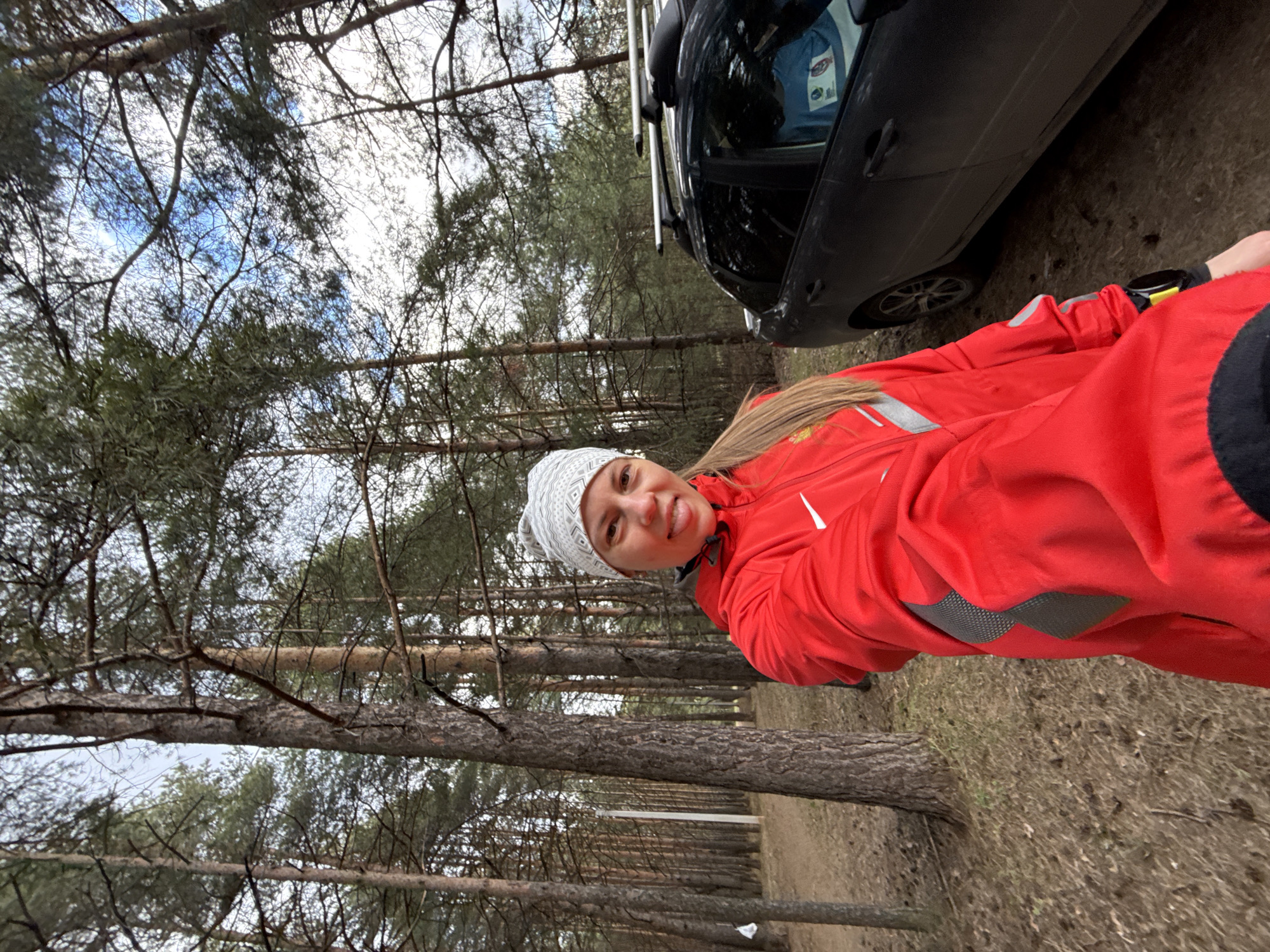 A woman with long blonde hair, wearing a bright red athletic jacket with a white Nike swoosh logo on the right shoulder and a white patterned beanie, is partially visible in the foreground of this outdoor image. She is smiling cheerfully and sticking her tongue out playfully, looking towards the camera. Gray textured patches are visible on the side of her red jacket.
Behind her, to the right, a dark-colored car, possibly a sedan or SUV, with a roof rack, is parked on a dirt surface. A light blue item with an indiscernible logo or text is visible through the car's window. The setting is a dense pine forest, with numerous tall tree trunks visible throughout the background. The ground is covered with dry leaves and sparse, dry grass, suggesting a colder season or dry conditions. A dirt path can be seen stretching into the distance among the trees.
The sky, visible through the tree canopy, appears partly cloudy or overcast, indicating daytime. The overall atmosphere is casual and outdoorsy. The image captures a moment of lightheartedness in a natural, wooded environment near a parked vehicle, consistent with a stop in a forested area outside Chaikovsky, Russia.