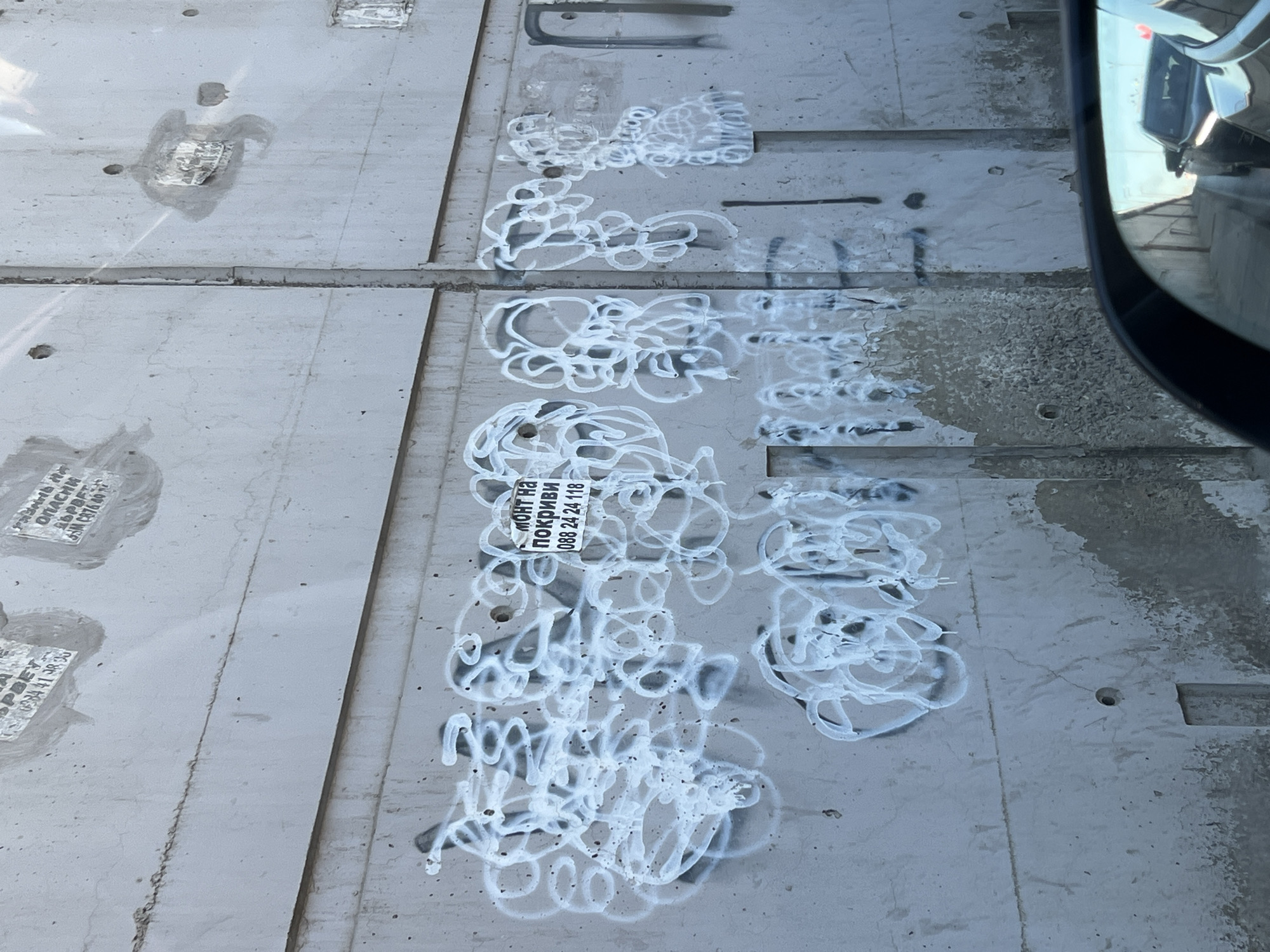 The image depicts a concrete surface in Sofia, Bulgaria. The surface is composed of multiple rectangular slabs. The slabs are visibly marked with white graffiti in a looping, abstract style. A small, rectangular sticker is affixed to one of the slabs, bearing the text "МОНТ НА ПОКРИВИ 088 24 24 118." The sticker appears to be partially obscured by the white graffiti. The slabs are separated by narrow gaps, and some are marred by stains and debris. The edge of a vehicle's side mirror is visible in the upper right corner of the image, reflecting part of the surroundings. Other markings, possibly construction labels or stamps, are visible on some of the slabs. The lighting suggests it is daytime. The environment seems to be an urban or industrial area, likely on a street or sidewalk.