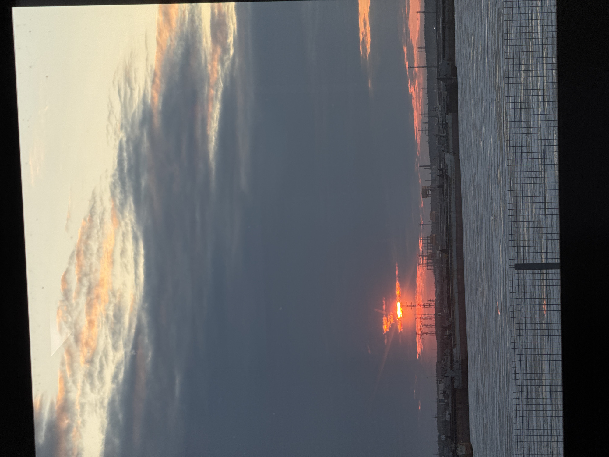 The image shows a sunset or sunrise scene, likely taken from a high vantage point, possibly a building overlooking a body of water. The sky is filled with clouds, some illuminated by the sun with an orange/red hue. The sun is visible, situated low on the horizon, suggesting either dawn or dusk. In the distance, there's a cityscape with buildings and structures including what appears to be power line towers, which can be common structures in industrial areas.
The right side of the frame includes a grid-like structure, which could be a window or a fence, providing a contrasting foreground element.
Given the city provided, Chaikovsky, Russia, is an industrial city situated in the Perm Krai region, the cityscape features may include structures affiliated with the city's industry. However, specific details about the location cannot be verified due to lacking visual cues.