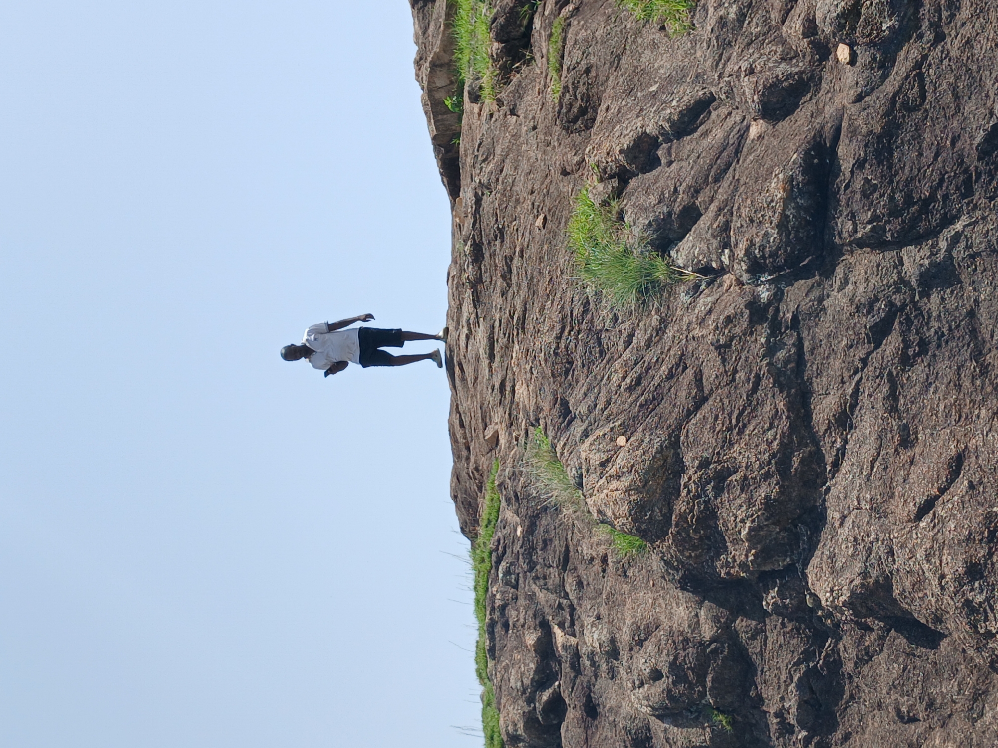 A man stands on a rocky incline under a clear sky. The man, wearing a light-colored short-sleeved shirt and dark shorts, holds a smartphone in his right hand. He is positioned towards the top of the frame, silhouetted against the bright sky. The ground beneath him is composed of rough, textured rock formations, with sparse patches of green grass growing in crevices. The overall scene suggests an outdoor, natural environment. The location is specified as Kwang, Nigeria.