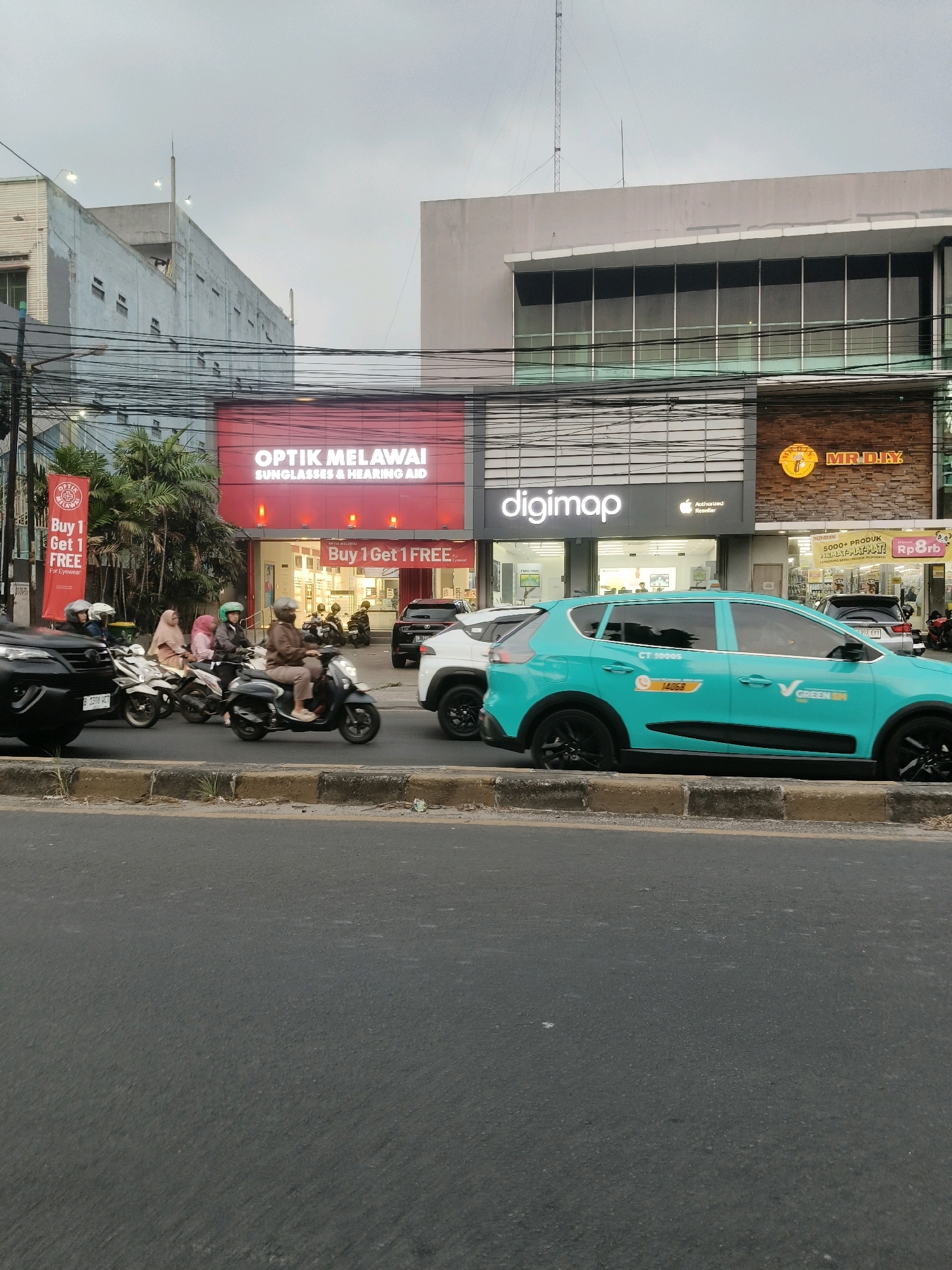 The image captures a street scene in South Tangerang, Indonesia, likely during daylight with overcast skies. In the foreground, a dark gray asphalt road with a concrete curb is visible. Behind the curb, several motorbikes and cars are present, suggesting moderate traffic.
The middle ground features a row of storefronts. On the left, "OPTIK MELAWAI" is prominently displayed with "SUNGLASSES & HEARING AID" written below it. A red banner for "Buy 1 Get 1 FREE" is hung vertically next to the entrance. Adjacent to Optik Melawai is "digimap," an Apple authorized reseller, with a clear display window. To the right of digimap is "MR. DIY," a retail store, identifiable by its brick facade and signage.
Several individuals are riding motorbikes, some wearing helmets, navigating the street. The people on the motorbikes appear to be dressed in casual clothing. A bright teal car with "GREEN SM" and a phone number "14068" on its side is parked near the curb, likely a ride-sharing vehicle.
The background consists of buildings with multiple stories, some appearing to be commercial or office spaces, with windows and electrical wires crisscrossing the scene. The overall impression is that of a busy urban commercial area.