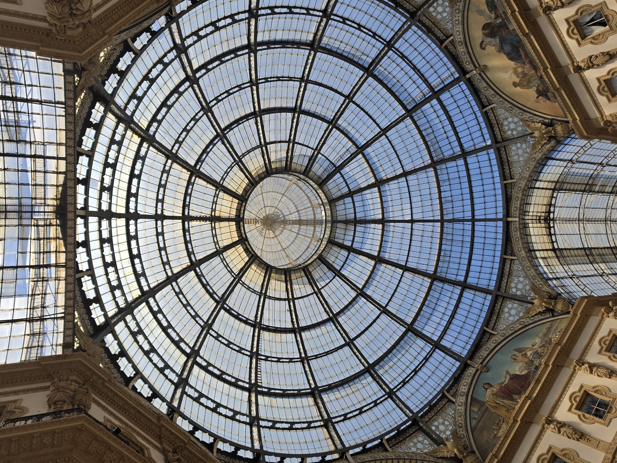 The image shows the interior of the Galleria Vittorio Emanuele II in Milan, Italy, specifically looking upwards at its magnificent glass and iron dome. The dome is octagonal in shape and is composed of intricate metalwork supporting a grid of glass panes. Sunlight streams through the glass, illuminating the interior. Below the dome, ornate architectural details are visible, including arched frescoes depicting mythological or allegorical scenes, sculptures, and decorative moldings. The overall impression is one of grandeur and architectural brilliance. There are no people visible in the image.