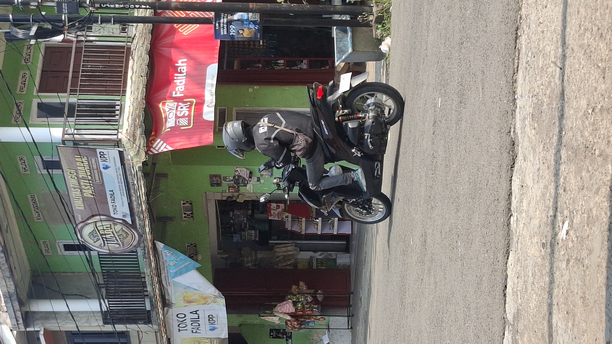 In Cilaku, Indonesia, a person wearing a dark helmet, black jacket, and blue jeans rides a dark-colored scooter. The rider is looking down at a smartphone held in their left hand. The scooter has a brown bag slung across its handlebars. The scene is set in front of a green building with a balcony. A red banner hangs from the building, displaying "TOKO SRC Fadilah" in white and red lettering, and a slogan "Murani Pesan Antar" which means "Order Delivery". Another banner above the red one reads "KUALITAS TINGGI PASTI JUARA!" with the logo "JUARA" and "TOKO FADILA KPP". A smaller white banner with "TOKO FADILA KPP" is also visible. The building appears to be a shop or store, with goods displayed inside. To the right of the scooter, a black pole with wires and a blue and yellow advertisement is visible. The ground is paved with asphalt.