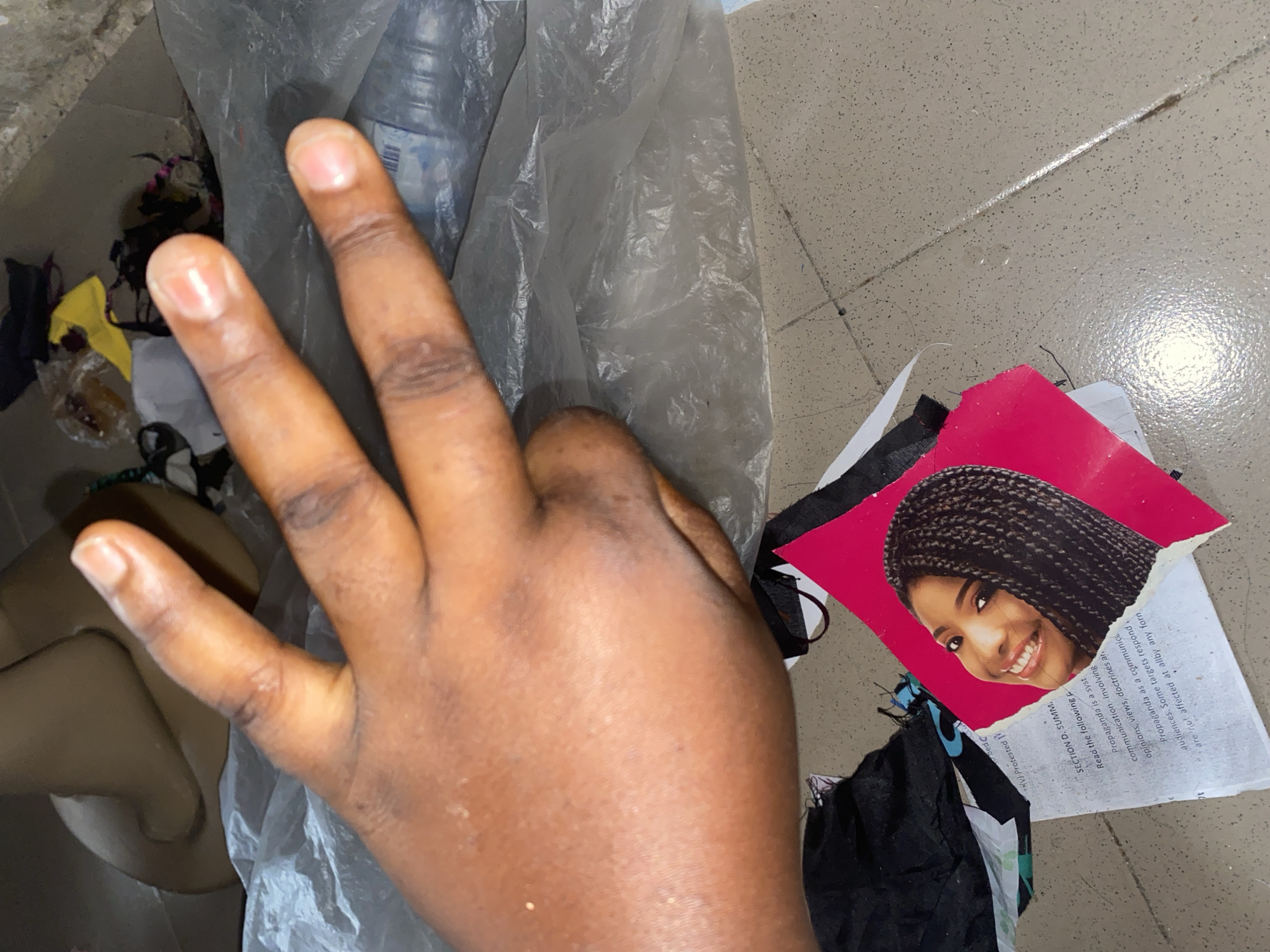A close-up, low-angle shot shows a person's right hand, fingers extended upwards in a "peace" or "victory" sign, with the thumb and index and middle fingers visible and separated. The hand is positioned in the foreground, with a blurry, pale brown, mannequin-like form partially visible behind it. The background is cluttered with various items, including crumpled clear plastic sheeting, black and yellow fabric scraps, and what appears to be a torn piece of paper with an image of a smiling Black woman with braided hair on a pink background. Below the image and to the left, more text is visible on a white paper, with the words "SECTION D. SUMM." and "Read the following p" clearly legible. The scene appears to be on a speckled, grey tiled floor. The overall impression is of a disheveled or discarded collection of items, with the hand gesture offering a moment of focus amidst the chaos.