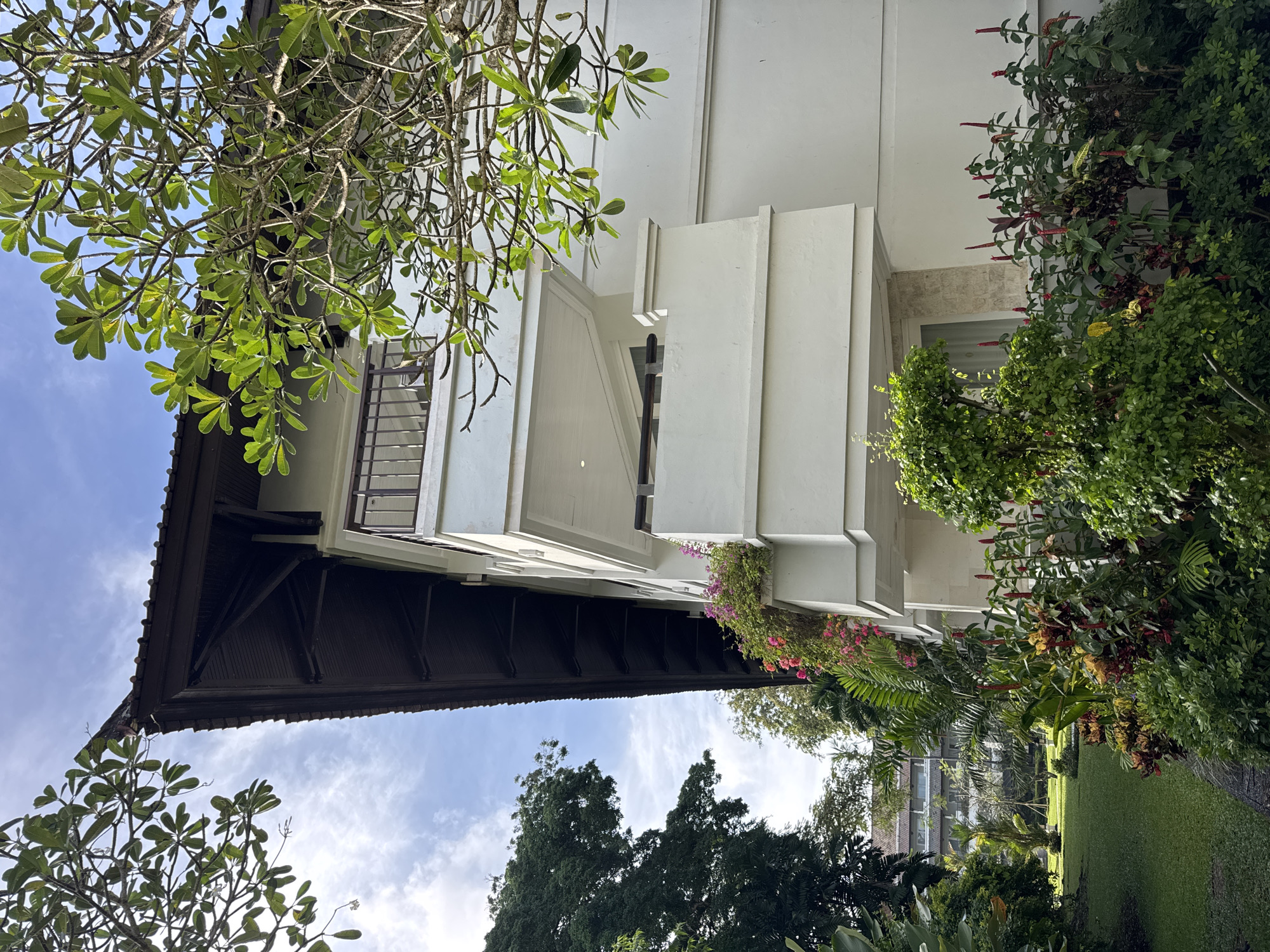 An outdoor scene captures a white, multi-story building with a dark, traditional roof, enveloped by abundant tropical vegetation under a bright sky. The building features distinct architectural details, including protruding sections, balconies, and windows, some fitted with decorative metal grates.
Lush green foliage dominates the foreground and midground. A large tree with vibrant green leaves and prominent branches, possibly a frangipani, frames the upper left, while dense shrubs and flowering plants with pink, purple, and red blossoms are integrated into the landscaping around the building. These plants create a vibrant, natural contrast against the white structure.
The sky is a clear blue with scattered white clouds, indicating daytime and fair weather conditions. Sunlight illuminates the scene, casting highlights on the leaves and the building's surfaces, contributing to a serene atmosphere. In the distance, beyond the immediate structures and trees, a well-maintained green lawn and other buildings suggest a larger resort or garden complex. No people are visible, and no specific activities are taking place. The tropical flora and architectural style are consistent with the location context of Kuta Selatan, Indonesia. There is no visible text in the image.