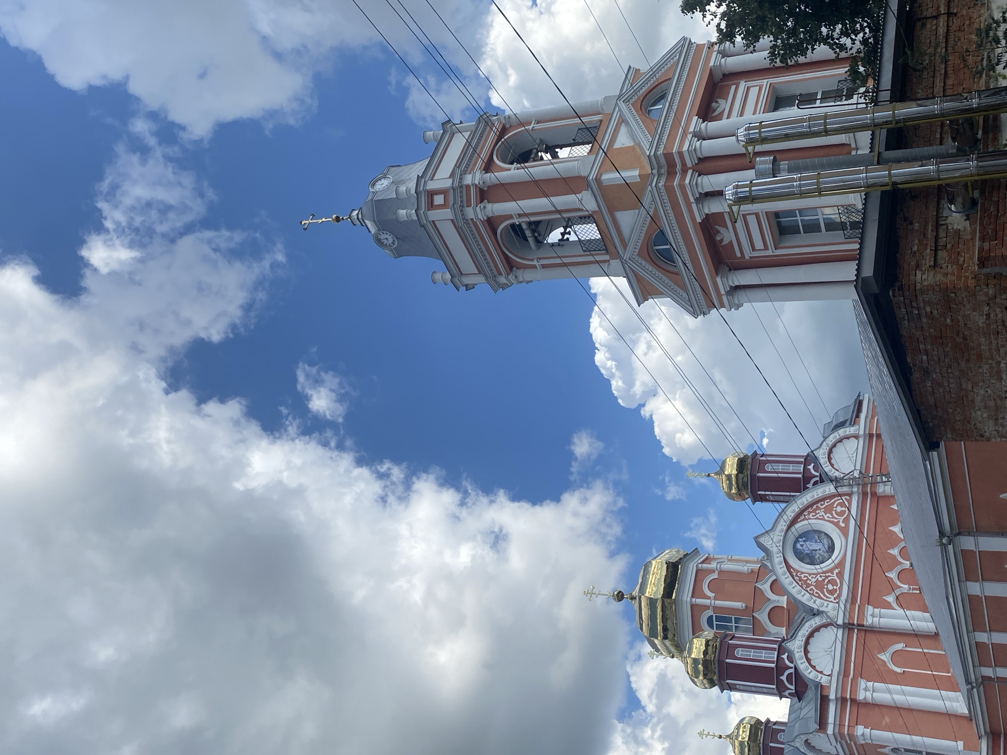 The image shows an exterior view of the Church of the Resurrection of Christ in Staraya Voskresenovka, Russia. The church features a prominent bell tower with a clock face and golden domes. The building is painted in shades of orange and white, with decorative white trim and columns. In the foreground, a brick wall and a metal roof partially obscure the lower portion of the church. The sky is blue with scattered white clouds. Overhead power lines are visible.