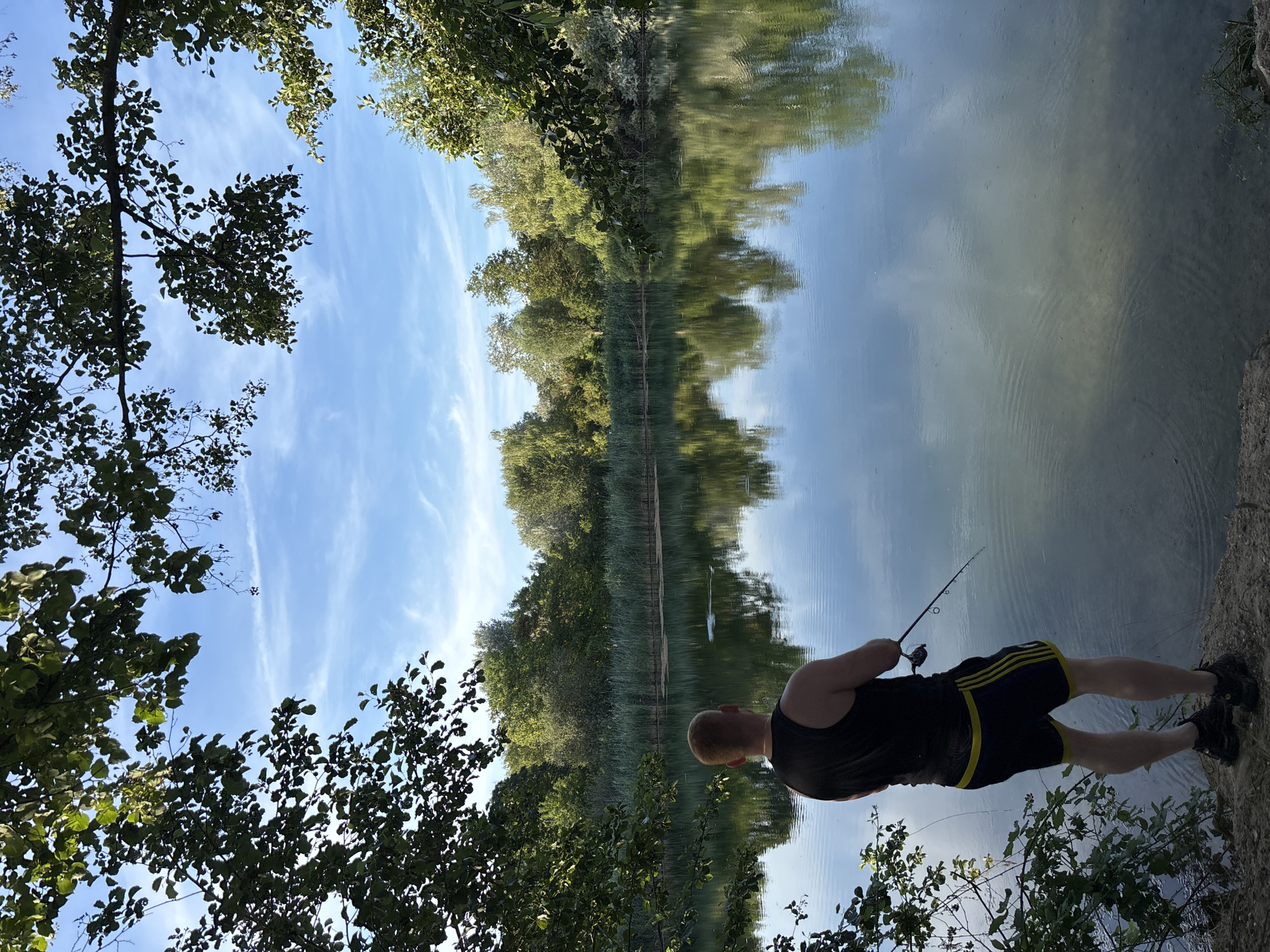 A man with red hair, wearing a black tank top and dark athletic shorts with yellow stripes, stands on the bank of a calm body of water, fishing. The man is viewed from behind, facing the lake. He holds a fishing rod, with the line extending into the water. Trees and reeds line the opposite bank of the lake, their reflections clearly visible on the water's surface. The sky above is blue with scattered white clouds. The foreground shows the man's feet and the rocky, earthy bank he stands on, with some greenery to the left. The scene appears to be a natural outdoor setting, likely a lake or pond. This location is identified as a pond in the municipality of Ludwigshafen am Rhein, Germany.
