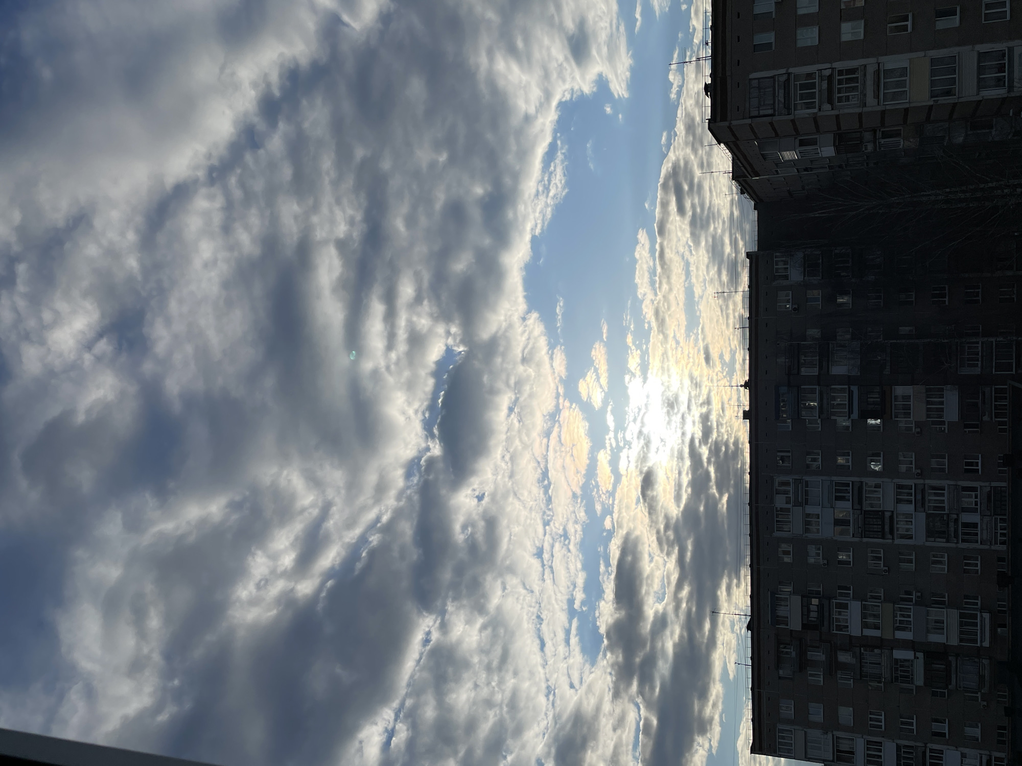 The image shows a daytime scene with a view of the sky and a building in what appears to be Izhevsk, Russia. The sky is filled with white and grey clouds, with some areas showing blue sky and the sun shining through. The sun's glare is prominent and located roughly at the center of the image. To the right is a tall, Soviet-era apartment building. The facade is grey and features many windows. There are antenna-like structures on the roof of the building. The image is oriented vertically, and the scene is shot from a low angle.