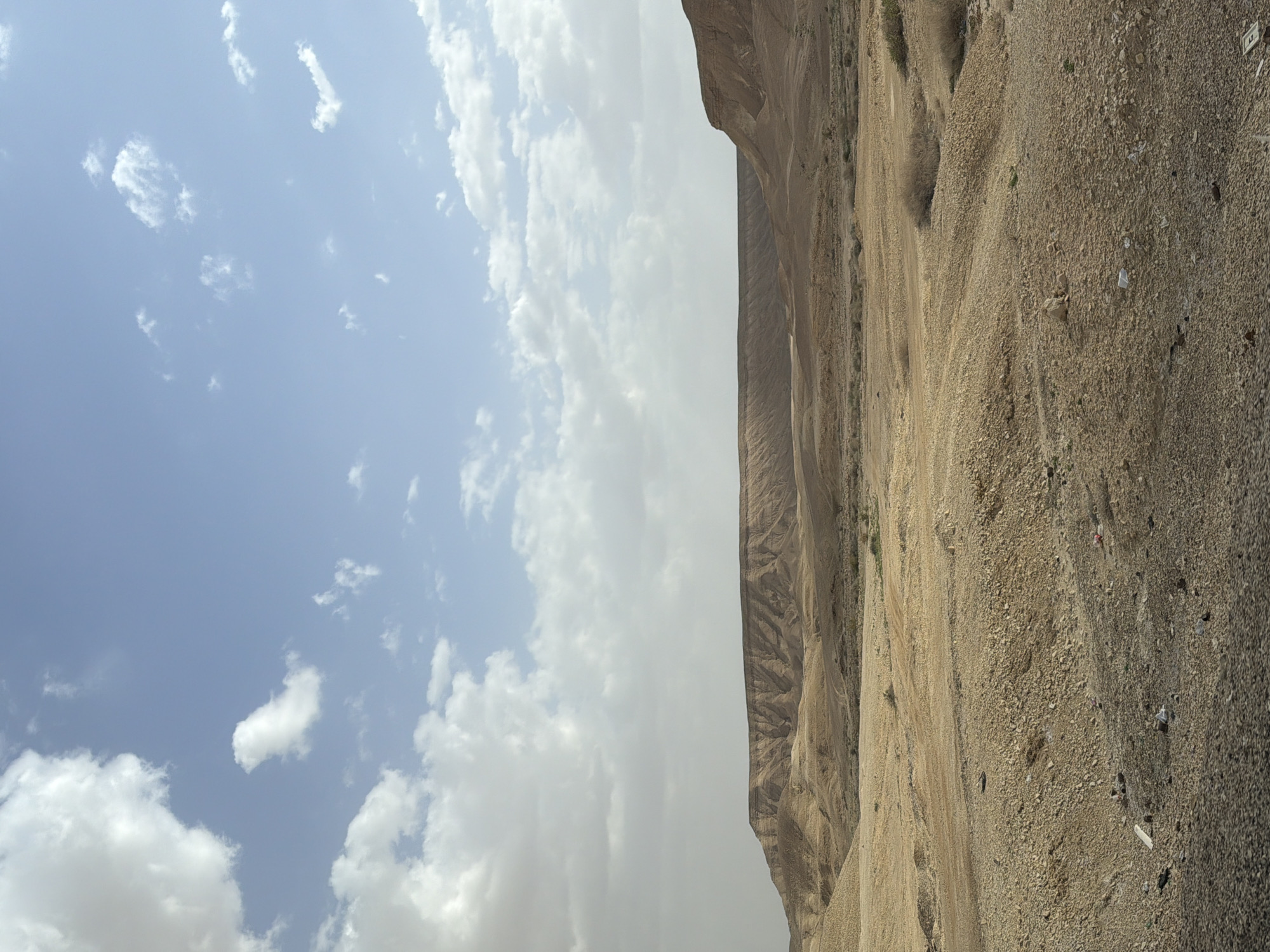 The image depicts a landscape in Tamar Regional Council, Israel. A mountain range occupies the right side of the frame, with its rocky, gravelly surface sloping downward. The sky takes up the left side, featuring a mix of blue sky and white clouds. The lighting suggests it is daytime. There are no people visible.