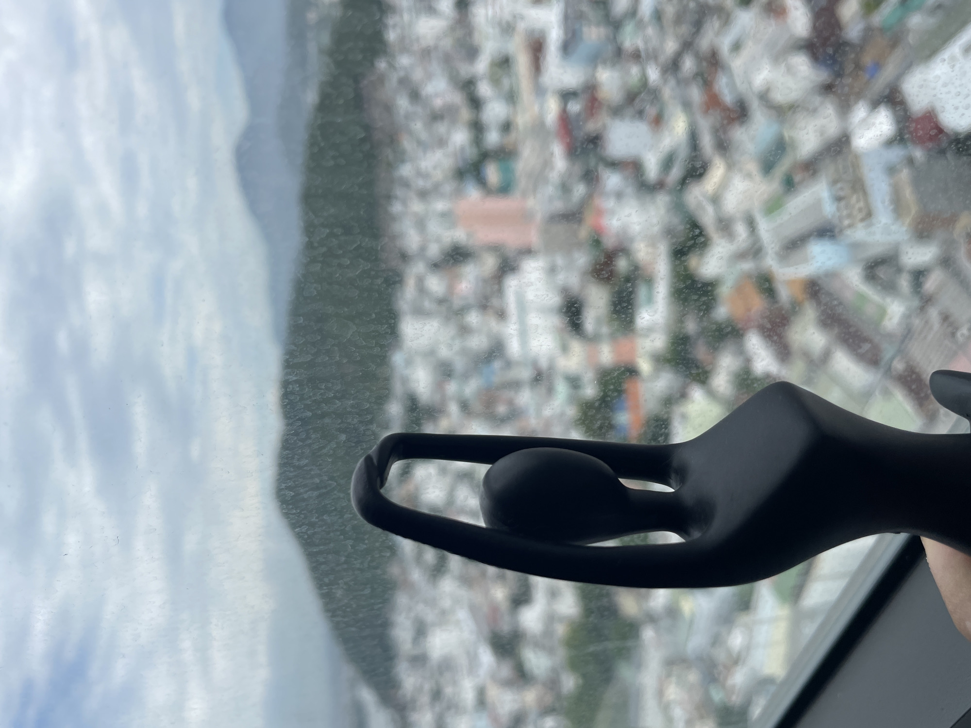 A black, abstract figurine of a dancer is held in front of a window overlooking a city and mountains. The figurine has its arms raised above its head, creating a smooth, curved arch. The city below is a blur of buildings, with some colorful roofs and structures visible. The windowpane has numerous water droplets, indicating it may be raining or the glass is wet. In the background, hazy mountains form the distant horizon under a cloudy sky. The overall impression is one of a contemplative moment, with the figurine acting as a focal point against the expansive, slightly obscured urban and natural landscape. Based on the visual cues such as the architecture and the lush, mountainous backdrop, this scene is likely located in Nha Trang, Vietnam.