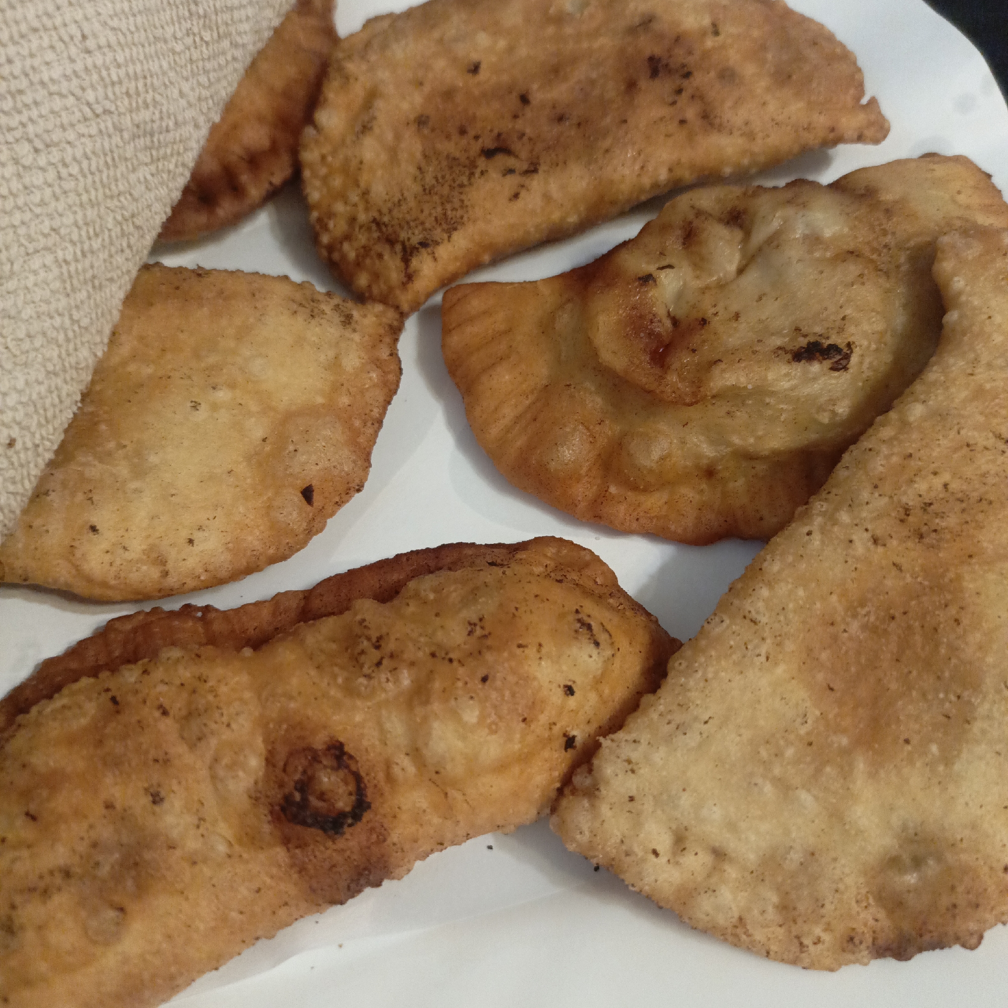 The image presents a close-up view of several golden-brown fried pastries, likely empanadas, turnovers, or similar savory or sweet fried dough pockets, arranged on a white plate. There are at least six individual pieces visible, exhibiting varying shapes, mostly crescent-like or semi-circular. The pastries have a distinct crispy texture, with some areas showing a deeper golden-brown hue and darker, almost charred, specks from frying.
A textured, light-colored fabric, possibly a beige or off-white towel or napkin, is partially visible on the left side of the frame, draping over one of the pastries. The white plate provides a clean and contrasting background for the food. The background behind the plate is dark and out of focus, suggesting an indoor setting, possibly a table. There are no people or specific activities depicted, beyond the presentation of the food. No text is visible in the image. The lighting highlights the fried texture and warm colors of the pastries.