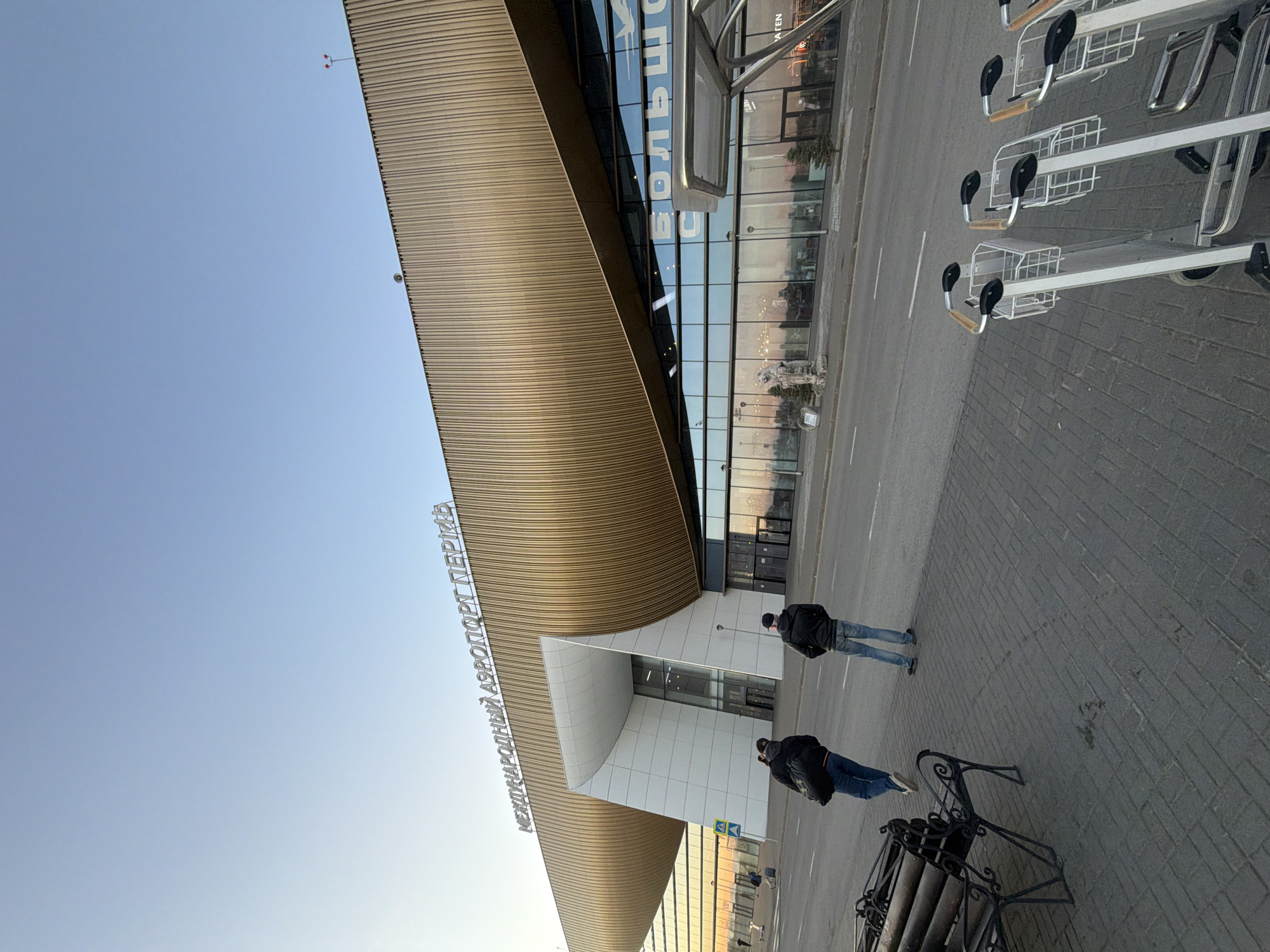 The image shows the front exterior of the Bolshoye Savino airport in Bolshoye Savino, Russia. The airport features a distinctive, curved facade with horizontal lines. The name of the airport is written in Cyrillic letters above the entrance. Two individuals are walking on the sidewalk in front of the building. A row of luggage carts is on the right side of the image. There is also a black metal bench. The sky is visible.