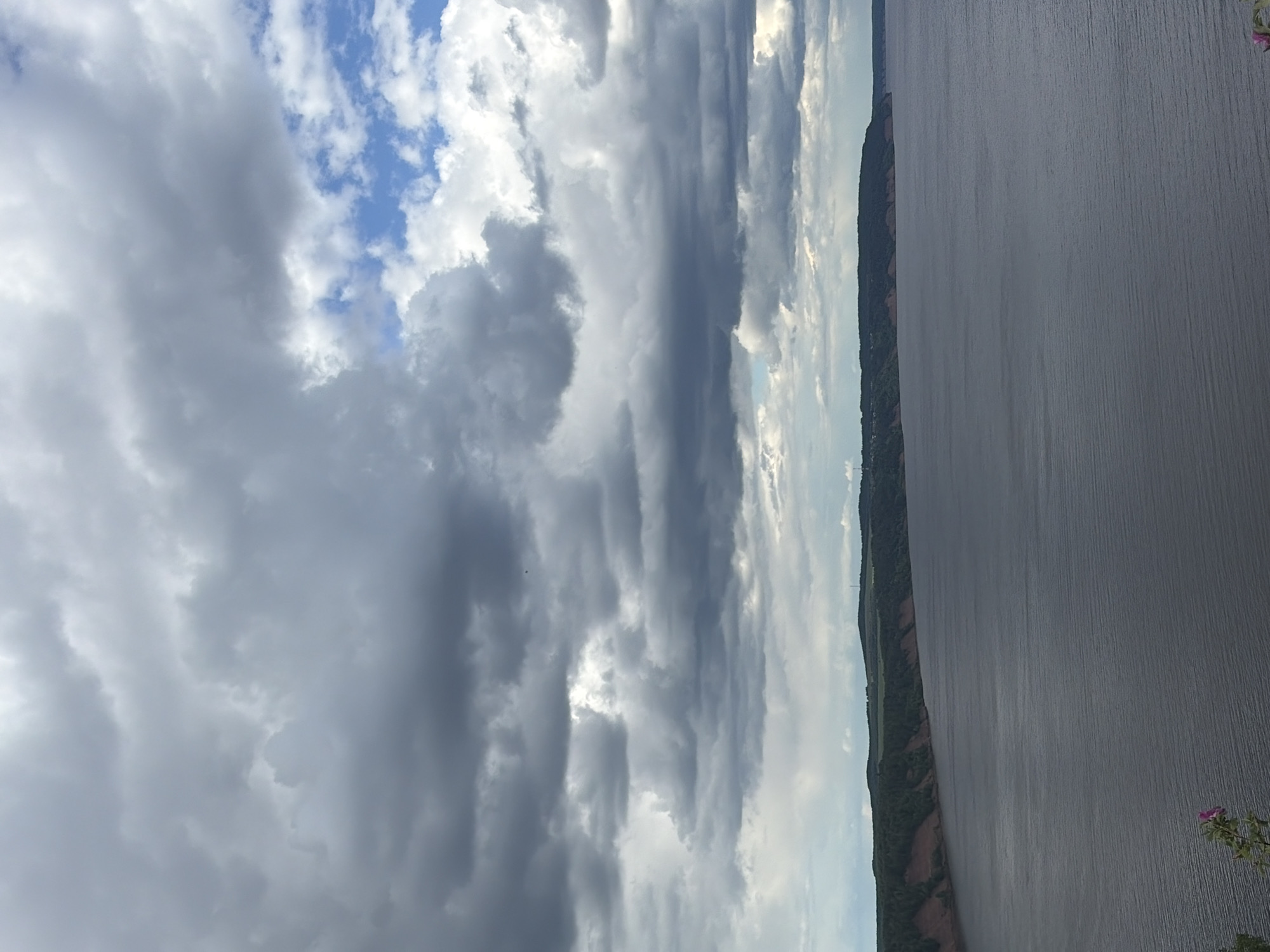 The image shows a landscape scene with a body of water on the right side, possibly a river or lake. The water surface is grey and appears calm. On the opposite shore, a strip of land is visible, featuring a mix of vegetation and some reddish-brown soil. Above the water and land, the sky dominates the view, filled with a variety of cloud formations. There are dark, heavy clouds as well as some patches of blue sky. The overall impression is of a somewhat overcast day. The provided location information, Костоваты, Russia, may be relevant in identifying the type of landscape and weather conditions typical for that region.