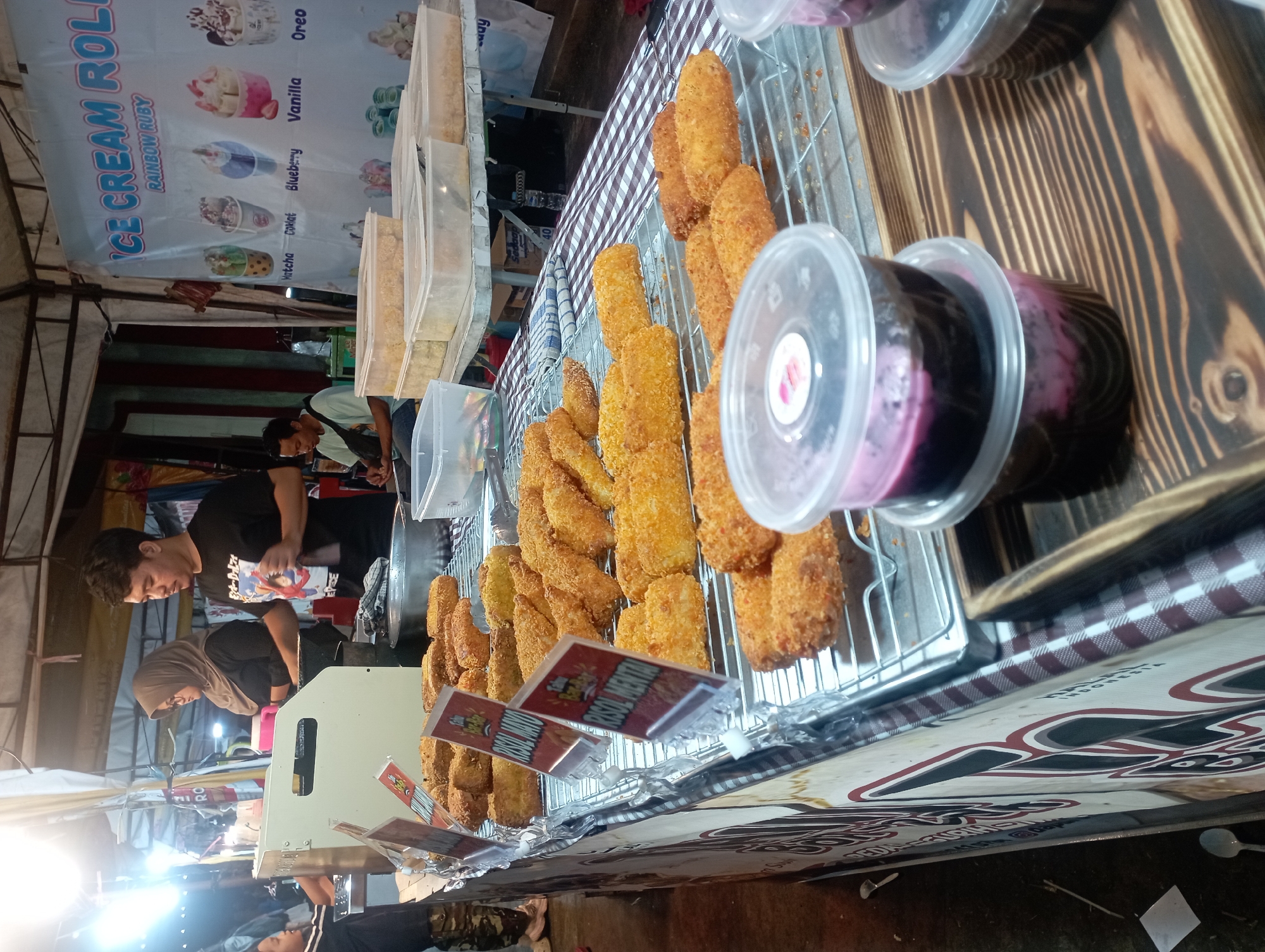 This image depicts a street food stall in Nganjuk, Indonesia, specializing in fried snacks and possibly ice cream. In the background, a woman wearing a hijab and glasses and a man in a black t-shirt are seen preparing food behind a counter. Another man is seated in the background, appearing to be a customer. The stall features a display of golden-brown fried items, likely risoles, arranged on metal racks. Several clear plastic containers filled with a dark purple mixture, possibly a dessert or drink, are also visible in the foreground. A banner advertising "ICE CREAM ROLL" with various flavor illustrations is prominently displayed. The stall is set up outdoors, likely at a night market or busy street, given the lighting and general atmosphere.