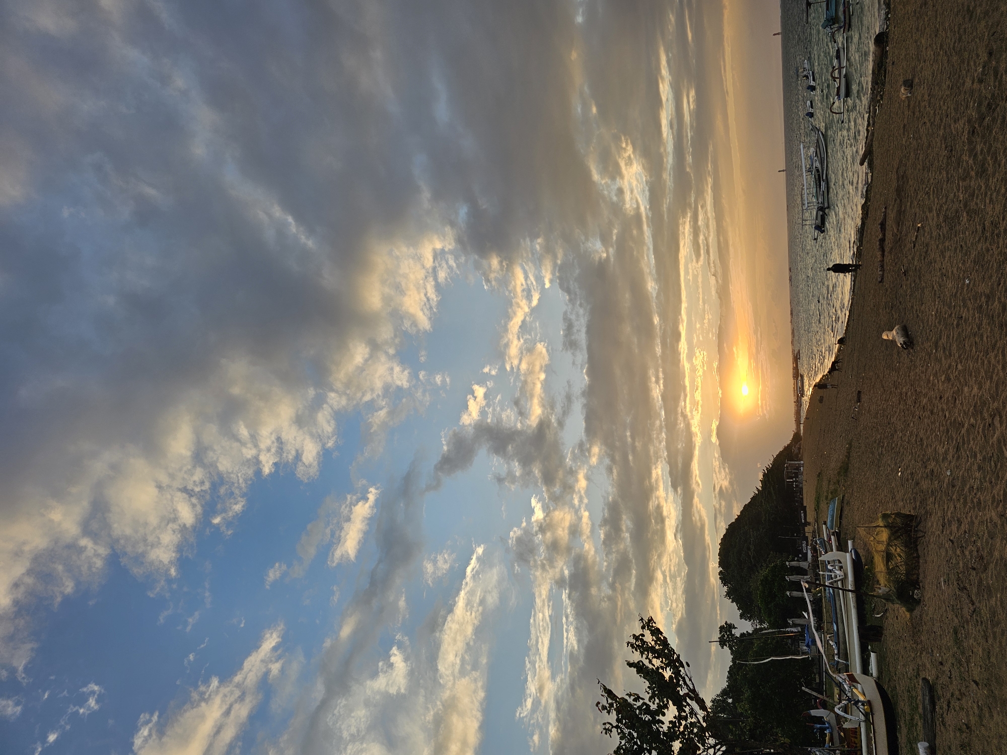 This is a wide-angle, eye-level, vertical photograph of a beach in Denpasar, Indonesia, during sunset. The sky is filled with a mix of blue and grey clouds, with the sun appearing as a bright orange orb low on the horizon, casting a warm glow. A line of trees and a hill covered in foliage forms the backdrop on the left side of the image, with several traditional outrigger boats, some with canopies, pulled up onto the sandy beach. A few people and a dog are visible on the beach and near the water's edge, where more boats are anchored. The water is calm with gentle waves. The sand is light brown and strewn with driftwood and some fishing nets. The scene is peaceful and serene.