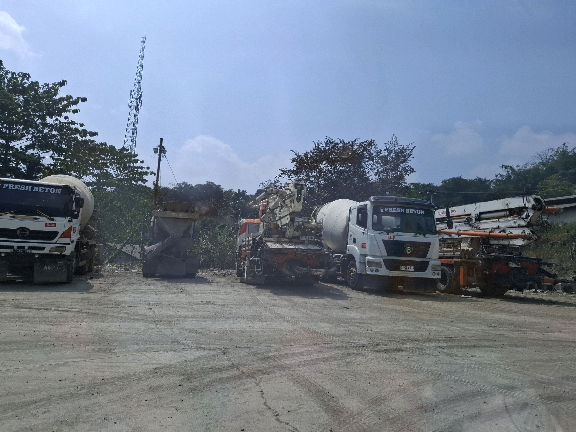 The image depicts an outdoor scene in the city of Cijangkar, Indonesia, likely a construction or industrial area. The foreground is a cracked, dusty concrete surface with tire tracks. In the midground, several large vehicles are parked. On the left is a white cement truck with "FRESH BETON" written on its side and a license plate "B 9247 BDB". Next to it is a piece of heavy equipment, possibly a loader, with a large bucket. To the right of the loader are two more vehicles. One is a concrete mixer truck with a white cab and "FRESH BETON" on its side, displaying the license plate "B 9158 TIA". Behind and to the right of this truck is a concrete pump truck. The background features a dense line of trees and vegetation, with a tall, slender telecommunications tower reaching into the clear blue sky. The overall impression is one of industrial activity.