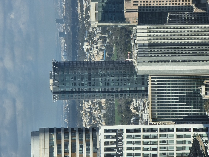 The image displays an elevated, wide-angle view of the Tel-Aviv cityscape, primarily centered on the Azrieli Center complex, under an overcast sky.
In the mid-ground, the prominent Azrieli Sarona Tower is visible as a tall, slender skyscraper with a dark blue glass curtain wall and a distinctive V-shaped white structural crown at its apex. To its immediate left, the Azrieli Circular Tower appears with a lighter-colored, rounded facade featuring stacked horizontal window bands. Partially visible in the lower-left of the frame, a white building segment displays "G GROUP" lettering on its upper section, consistent with the Azrieli Square Tower. In the lower-right foreground, the Azrieli Triangular Tower is present, characterized by its grey, vertically fluted concrete facade and numerous small, vertically aligned windows.
Surrounding these primary structures are multiple other urban buildings of varying heights, architectural styles, and material compositions, including glass and concrete facades. No individuals, vehicles, or specific actions are discernible within the frame. The urban environment extends into the distant background, showing a dense array of buildings interspersed with areas of green parkland, with additional, smaller high-rises visible on the distant horizon against the grey cloudy sky.