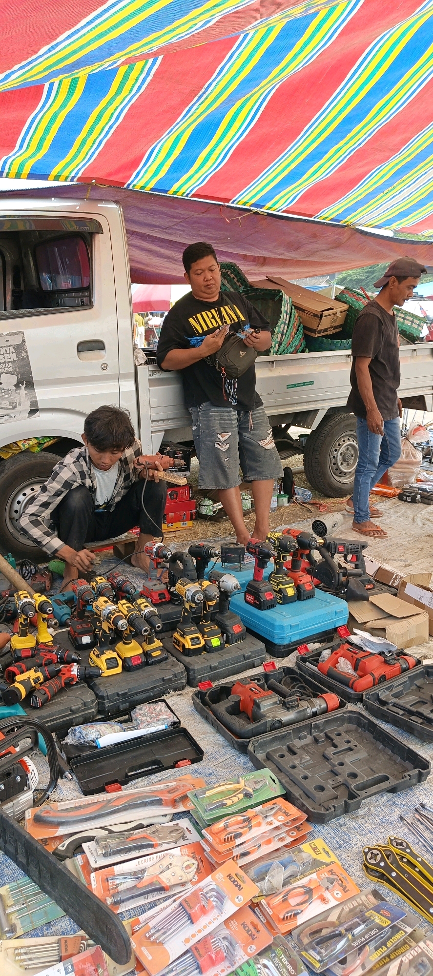 The image depicts an outdoor market scene in Jatinom, Indonesia, likely a Sunday morning market given the context of such events often occurring on weekends. The foreground is filled with various power tools, primarily cordless drills and impact drivers, neatly arranged on the ground in their cases. These tools are colorful, predominantly yellow, black, and red, suggesting they are new or in good condition. Further back, a pickup truck serves as a stall, with its bed loaded with what appears to be baskets or woven goods, and possibly more tools. Three men are present. One man, wearing a black t-shirt with a "Nirvana" logo and ripped denim shorts, is leaning against the truck, looking at his phone and holding what seems to be money or a small item in his hand. Another man, dressed in a plaid shirt and dark pants, is crouched down near the tools, appearing to be examining or holding a drill and a brush. The third man, wearing a dark t-shirt and blue jeans, is standing and looking towards the side, away from the camera. The market is covered by a striped tarp, with red, yellow, and blue stripes, providing shade. The ground is covered with a mix of fabric or tarp and loose earth. The overall impression is a bustling marketplace where tools are being sold.